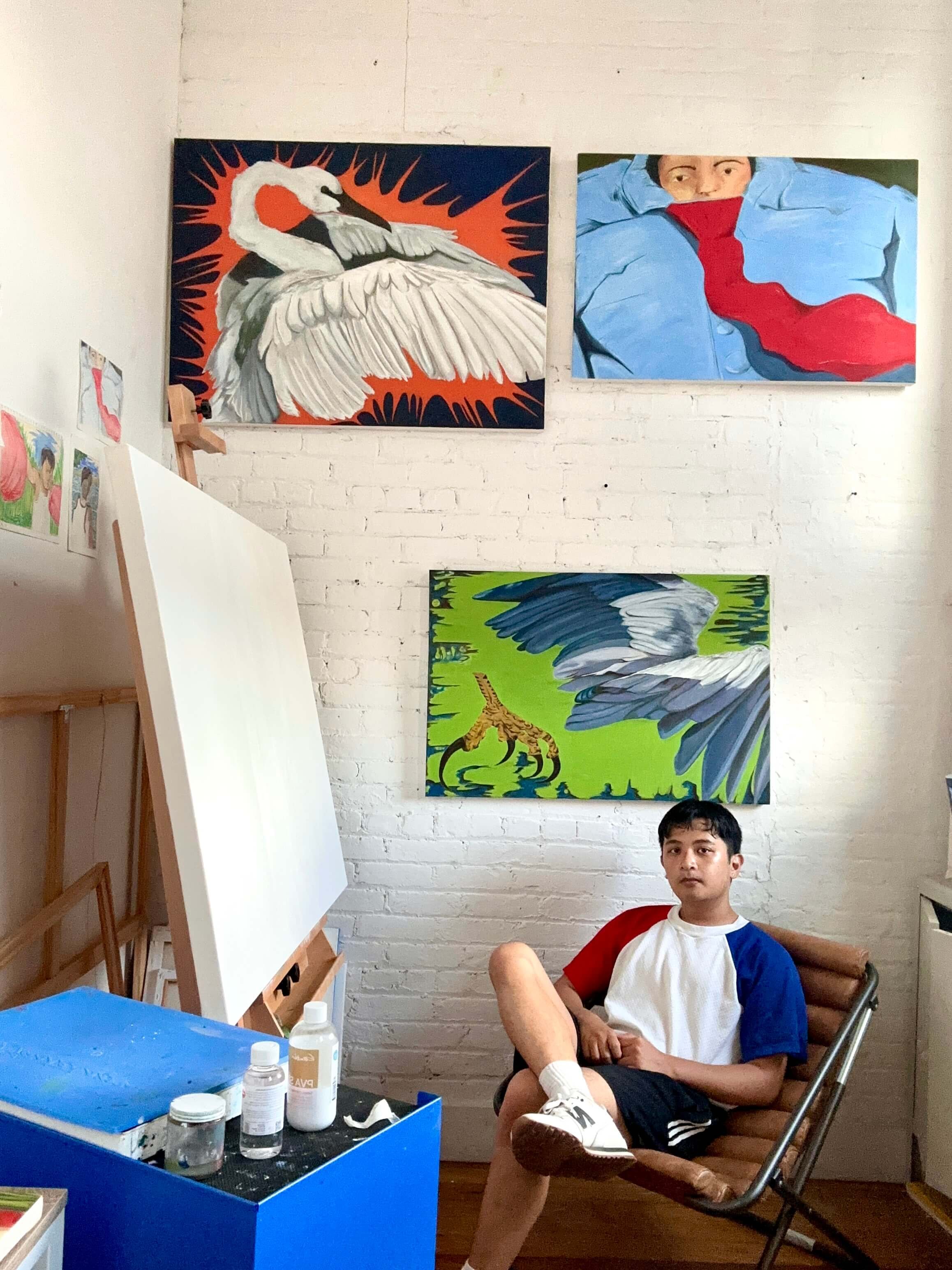 A boy sitting on a chair in an art studio with three paintings of animals and a person on the wall behind him.