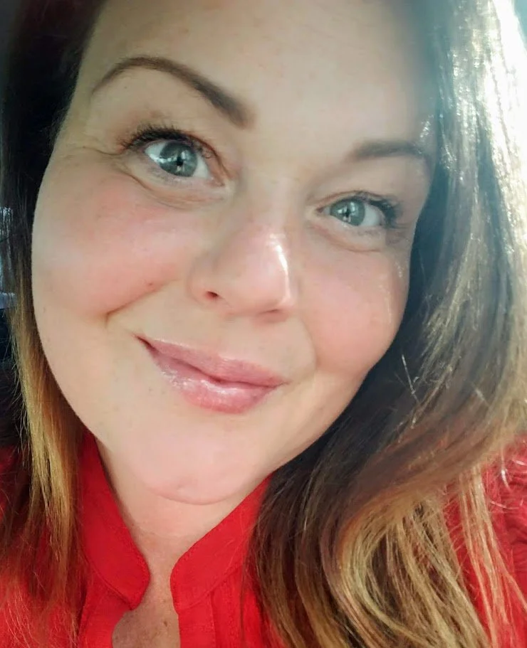 Close-up of a woman with long, wavy brown hair, blue eyes, and a bright smile, wearing a red top.