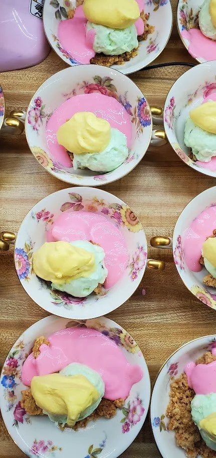 Several floral-patterned teacups filled with pink rose lassi and mint green pistachio fluff topped with yellow saffron whipped cream, placed on a wooden surface.