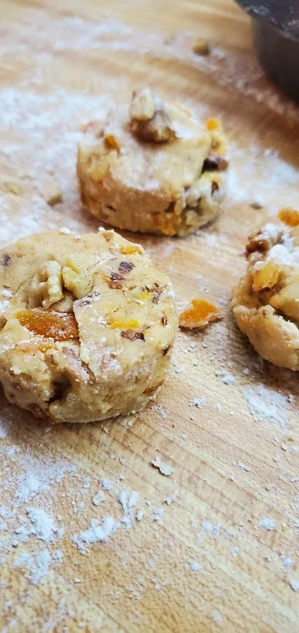 Unfinished scone dough with visible nuts and dried fruit on a floured wooden surface.