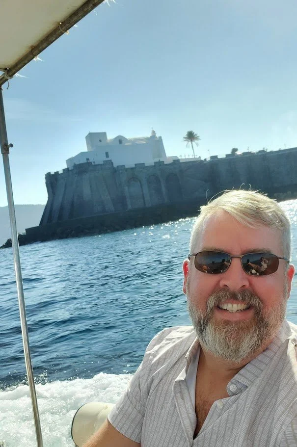A smiling man with sunglasses on a boat, with a historic fort and a palm tree in the background, on a sunny day.