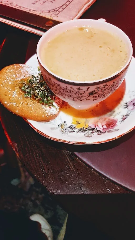 A cup of creamy tea and a tea cookie item topped with herbs on a floral plate