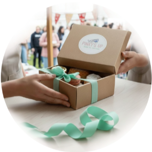 Person holding an open gift box with baked goods inside, tied with a teal ribbon, at an outdoor event with people in the background.