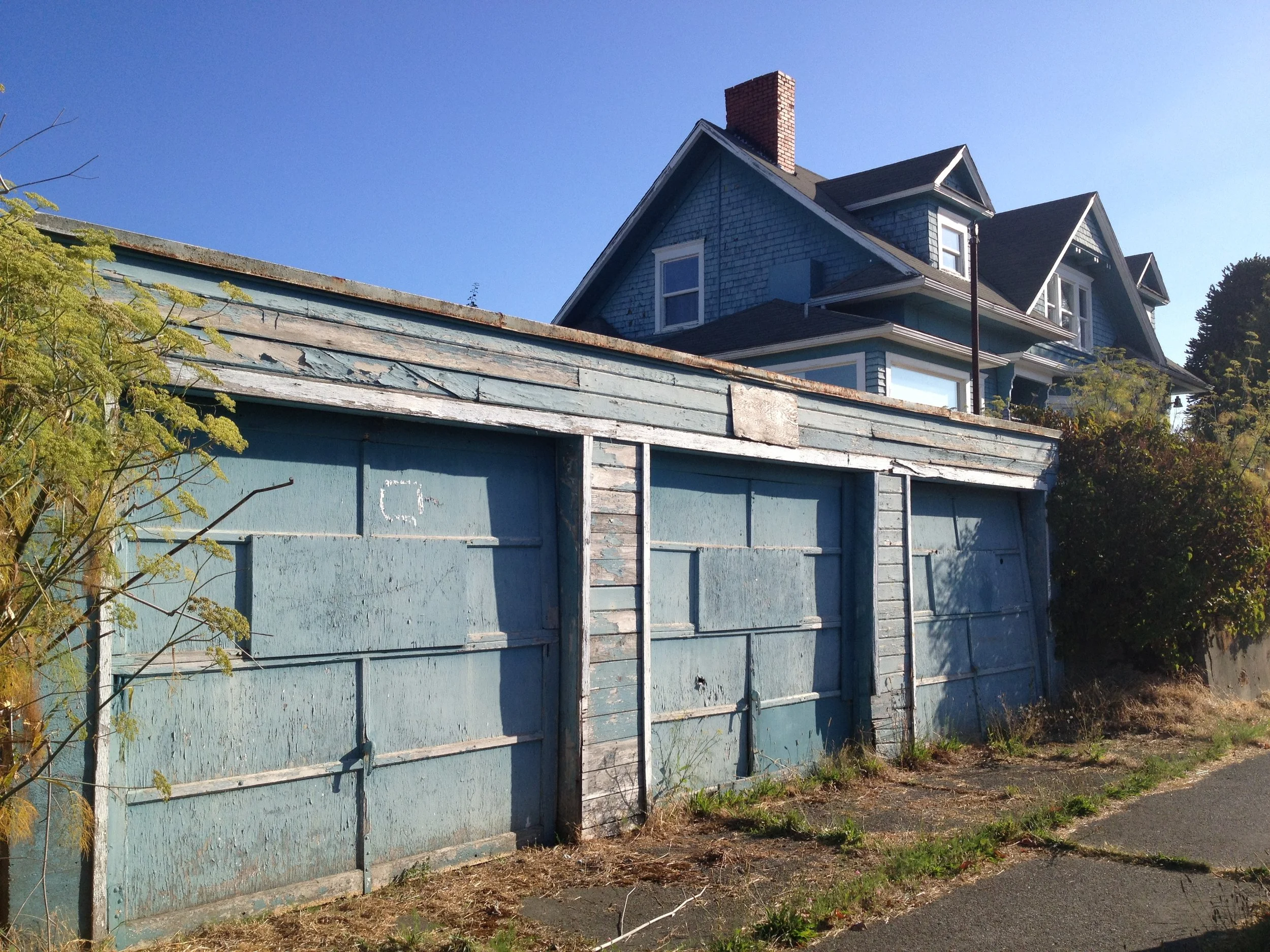 Old blue garage doors with peeling paint in front of a large blue Victorian-style house, clear sky, and some greenery.