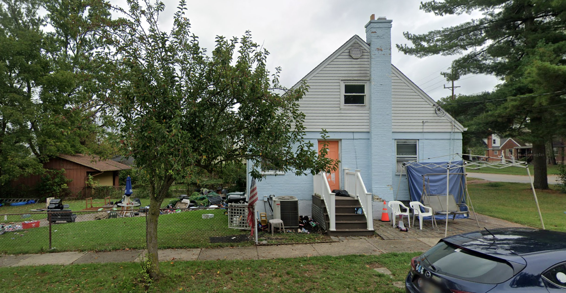 A blue house with a small front yard, a tree, and various outdoor furniture and objects, including a swing, chairs, and an umbrella.