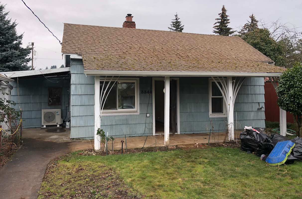 A small, light blue house with a yellowish-brown shingled roof and a brick chimney. The front porch has white decorative supports, and there is a small lawn in front. To the right, there are black trash bags and a blue plastic container on the ground. An air conditioning unit is on the left side of the house, and there are two windows and a door in the front.