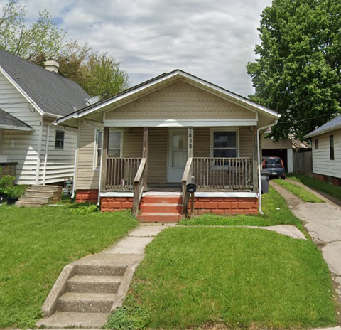 A small, single-story house with a front porch, white siding, and steps leading up to the entrance. The house has a green lawn and a sidewalk in front, with neighboring houses on either side and trees in the background.