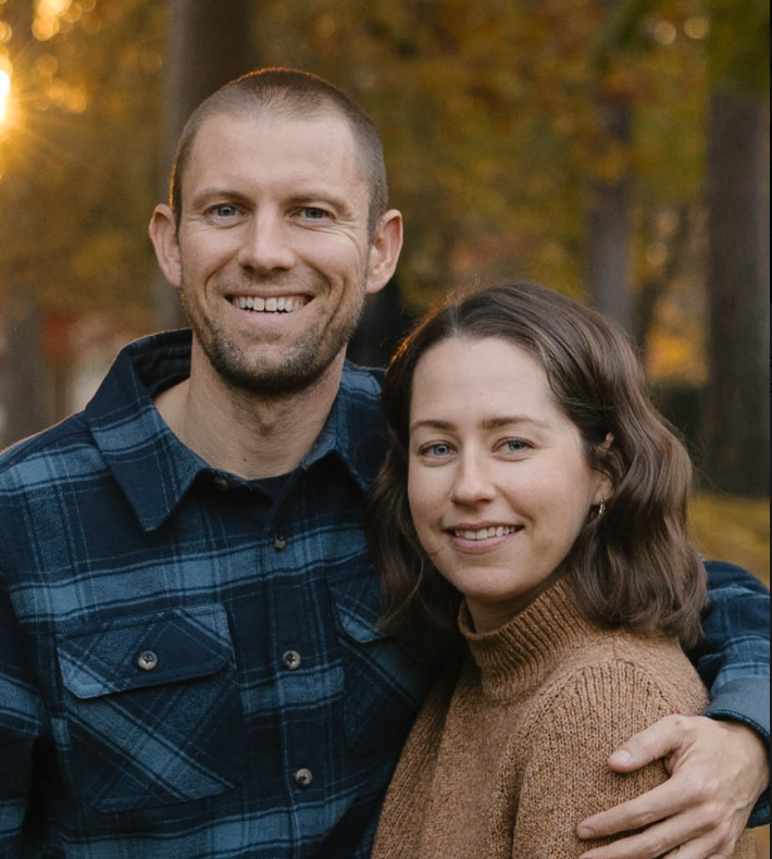 A smiling man and woman standing outdoors in a forest during autumn.