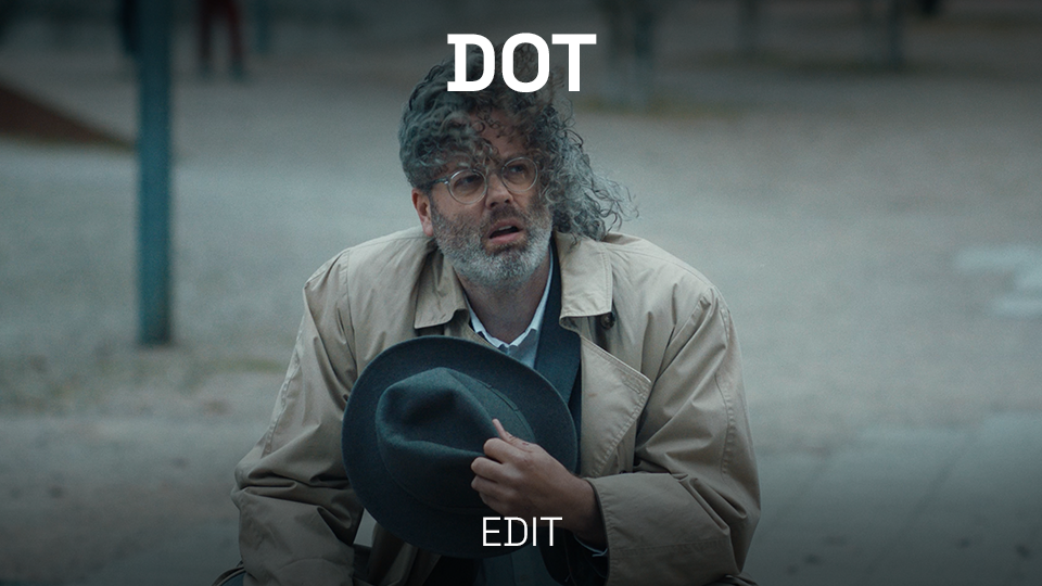 A man with curly hair, glasses, and gray beard holding a hat, appears to be in distress or confusion, outdoors in a park or street setting during daytime.