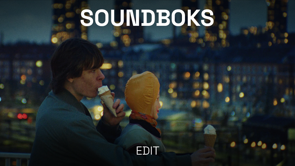 A young man and a child with a hood eating ice cream cones on an outdoor balcony at night, with blurred city lights in the background.