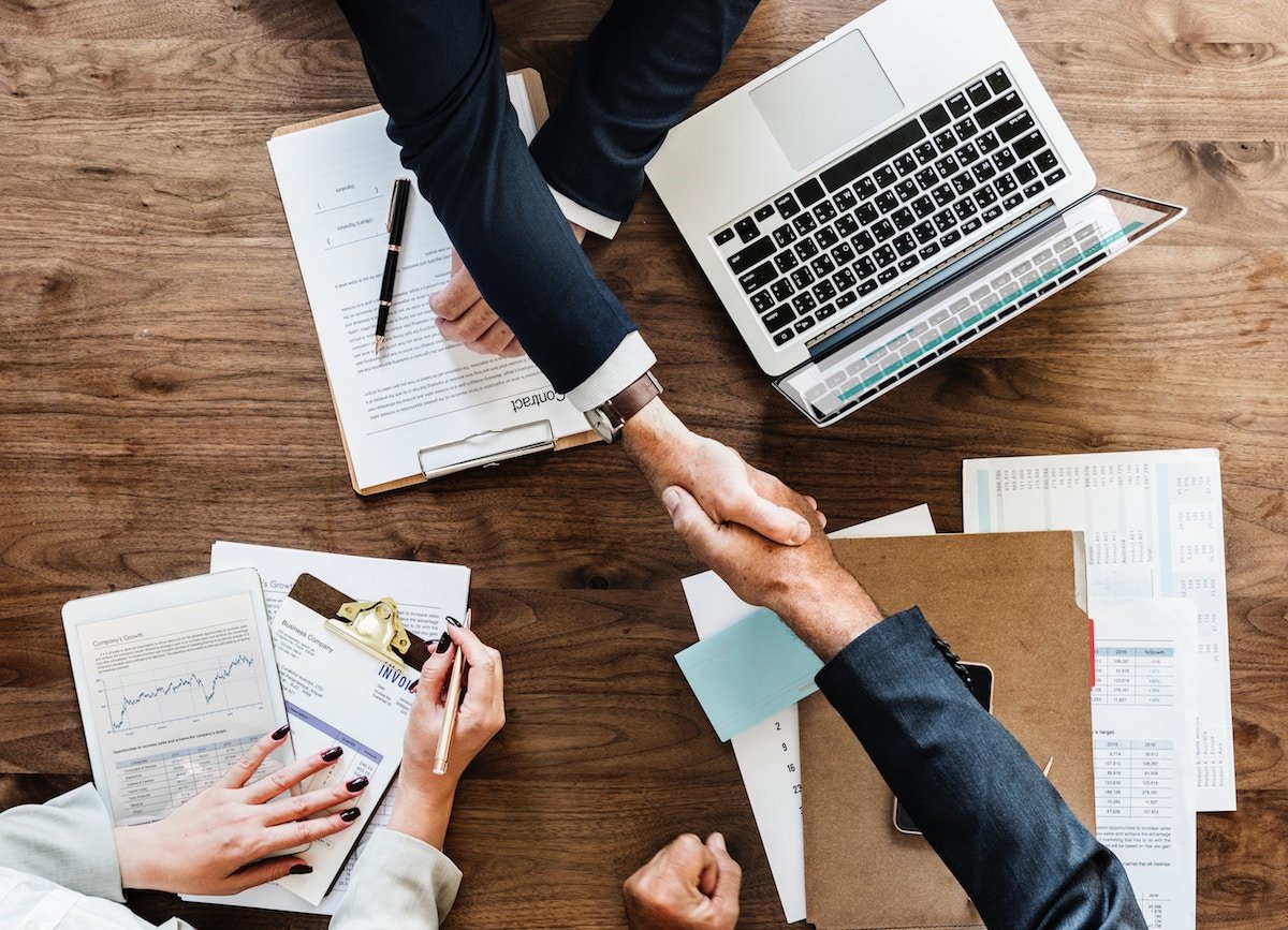 Two people shaking hands over a wooden desk covered with financial documents, charts, a laptop, and a clipboard.