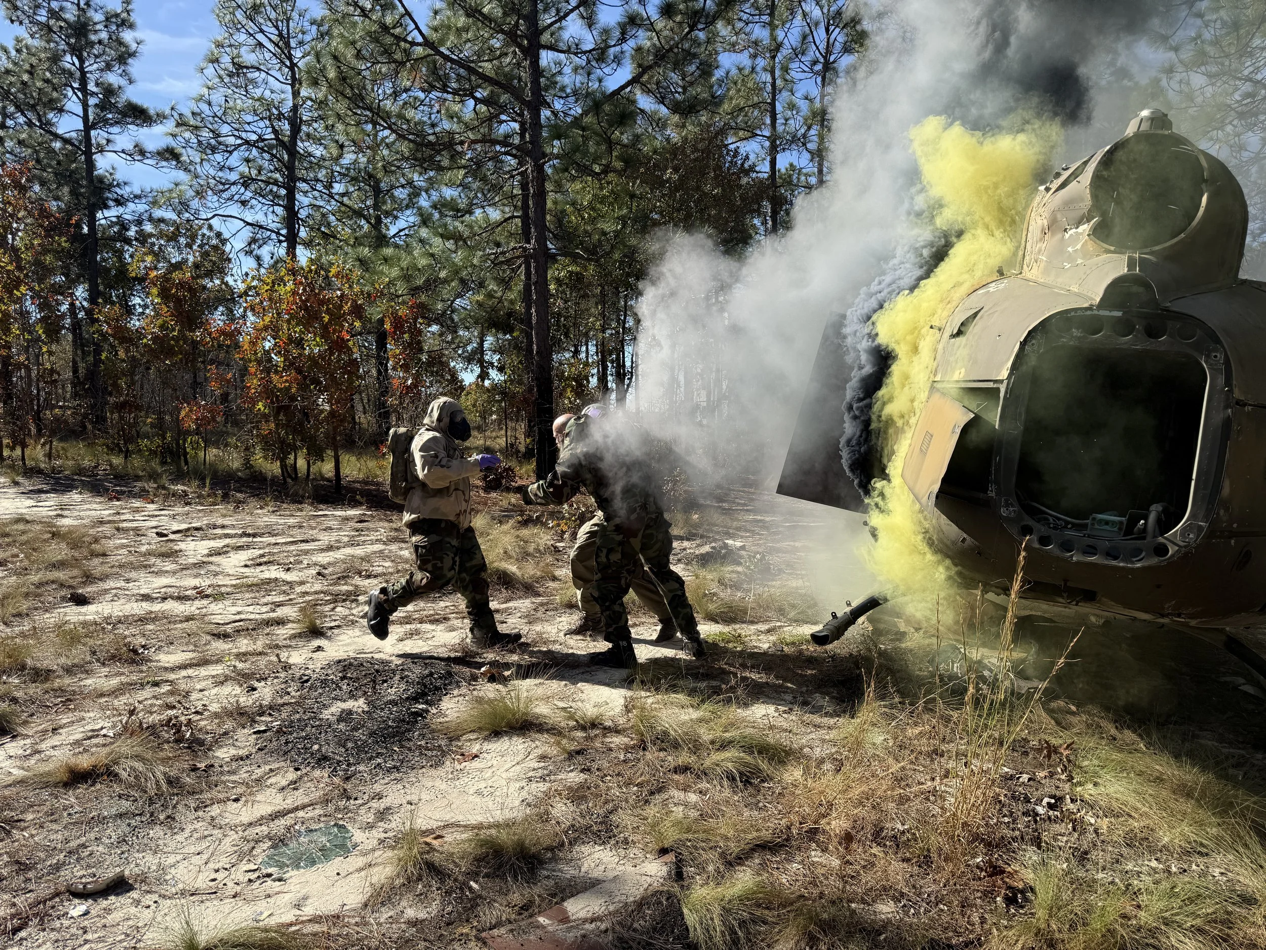 Two people dressed in military gear near a crashed helicopter with smoke and yellow foam, in a forested area.