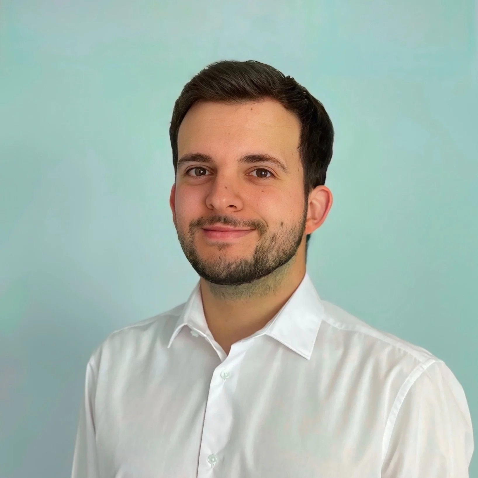 Portrait of a young man with dark hair and beard, smiling, wearing a white collared shirt, against a light green background.
