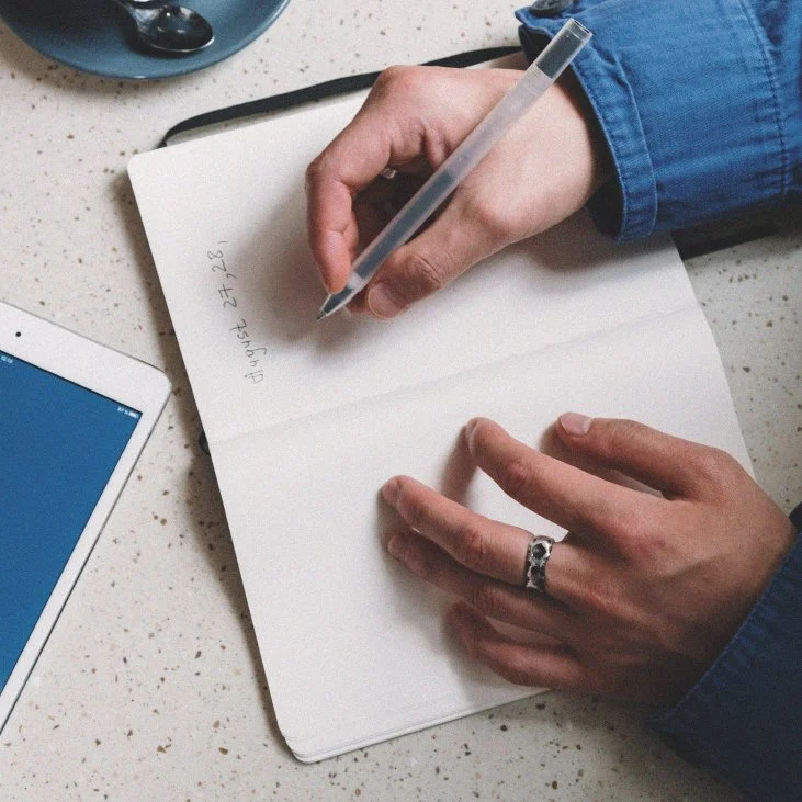 A person wearing a blue jacket writing in a notebook with a transparent pen. The notebook is on a speckled white surface, and a tablet and a black object are nearby.