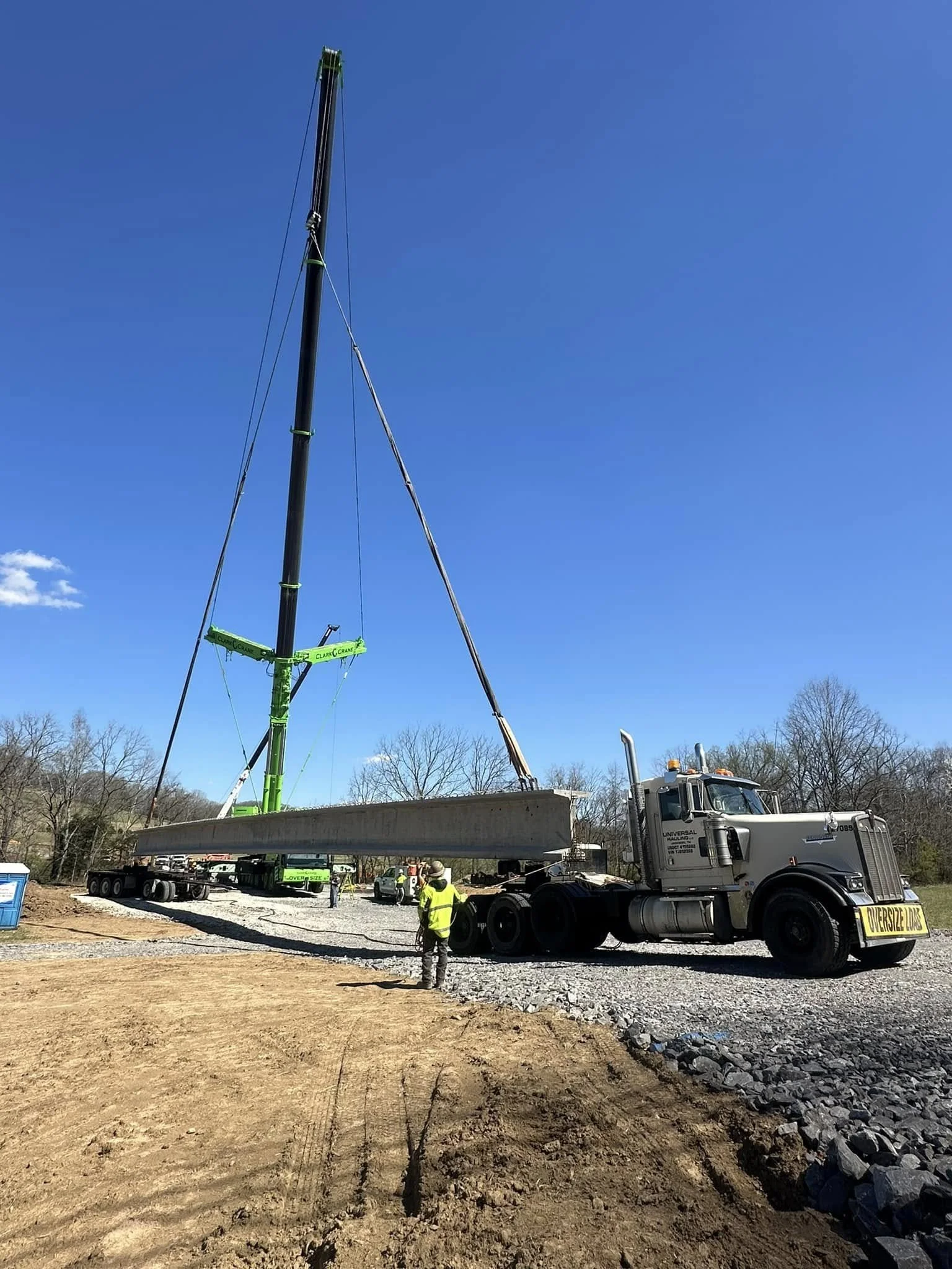 Construction workers use a crane to lift a large concrete beam onto a truck at a construction site under a clear blue sky.