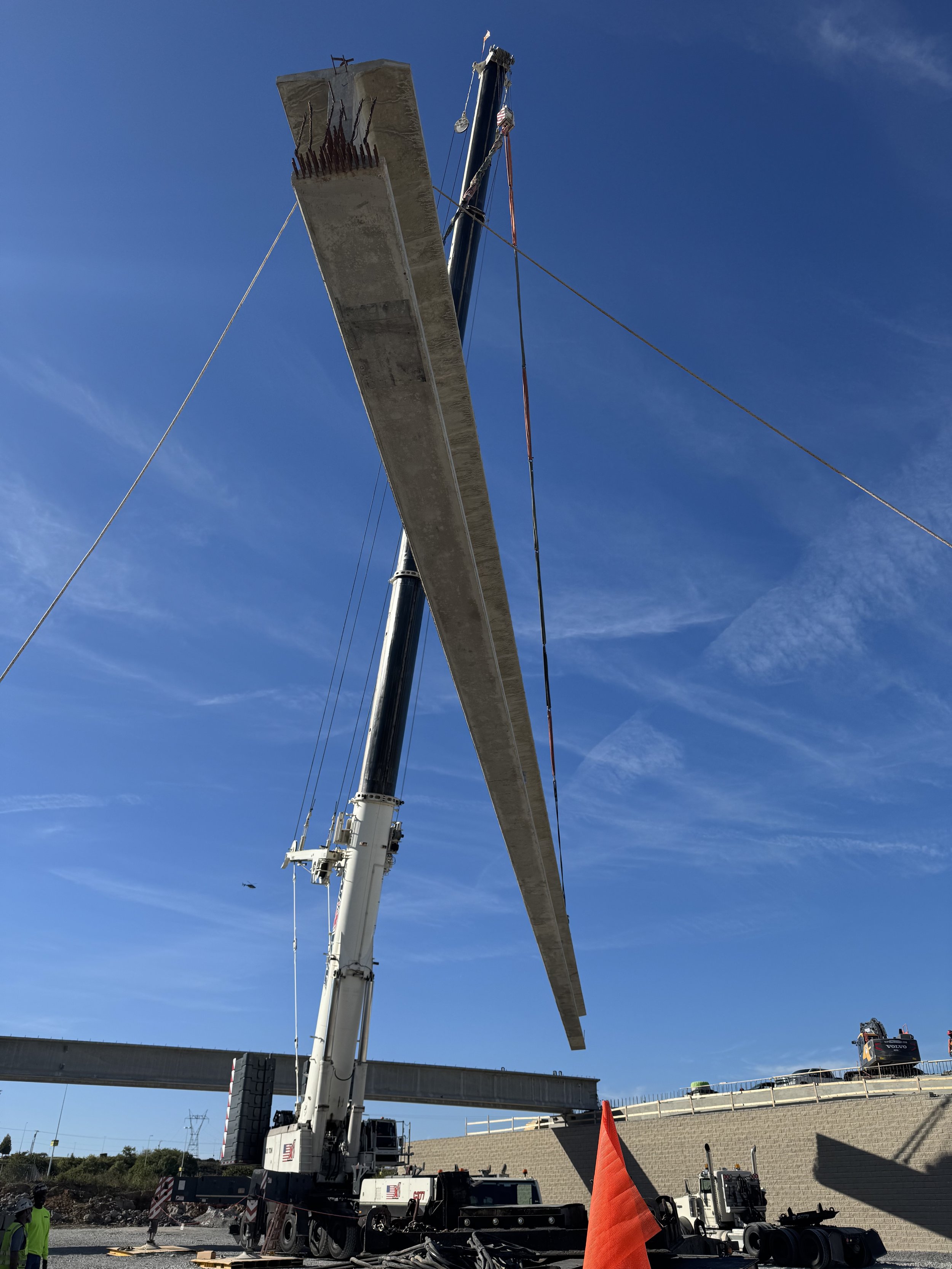 A large construction crane lifting a concrete bridge span into place during a road construction project, with a clear blue sky in the background.