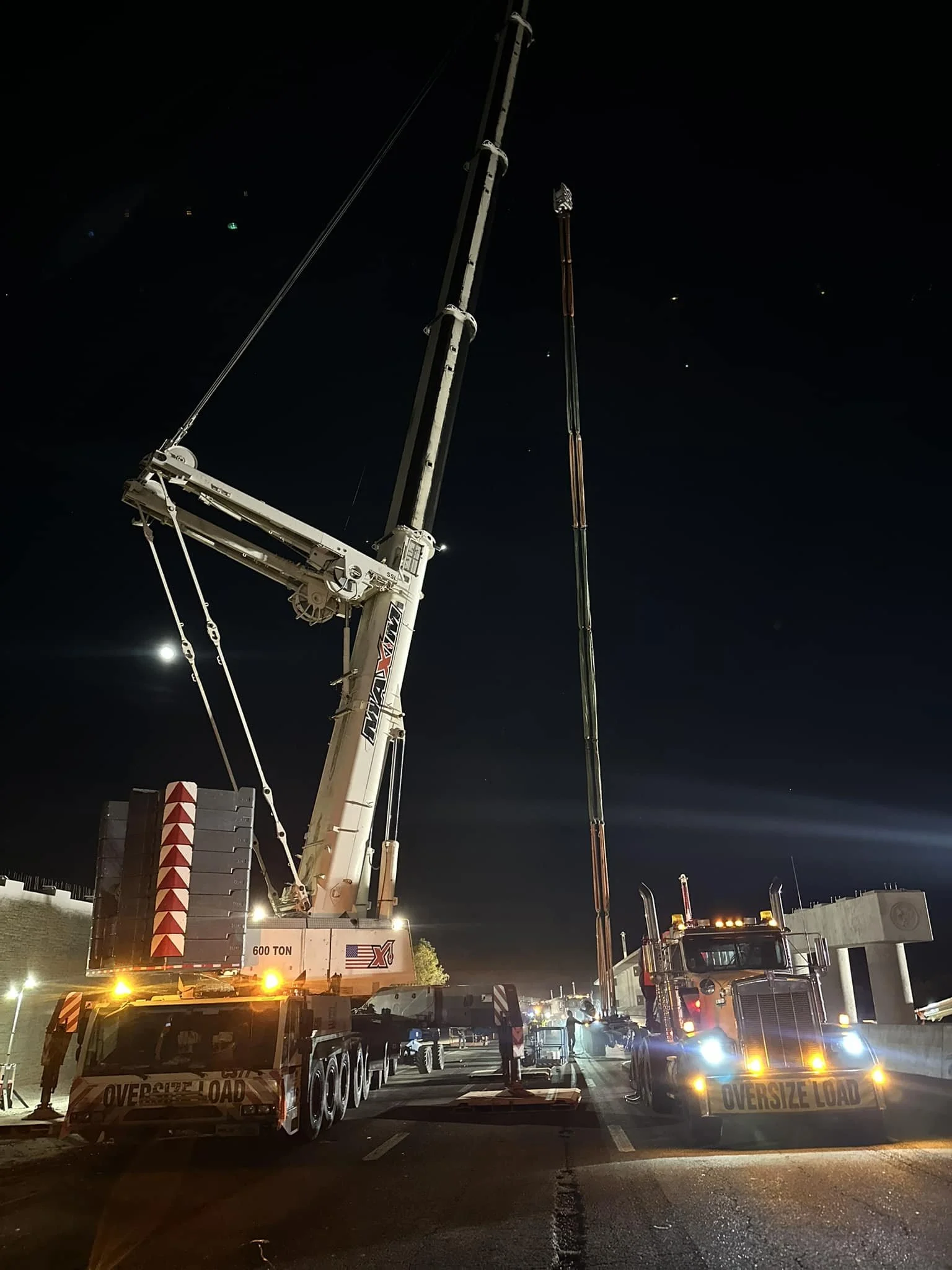 Nighttime construction scene with two large trucks, one with a crane and the other with an extendable boom, both marked with 'OVERSIZE LOAD'. The crane is lifting a long, tall structure over a highway, with workers present and traffic barriers in place.