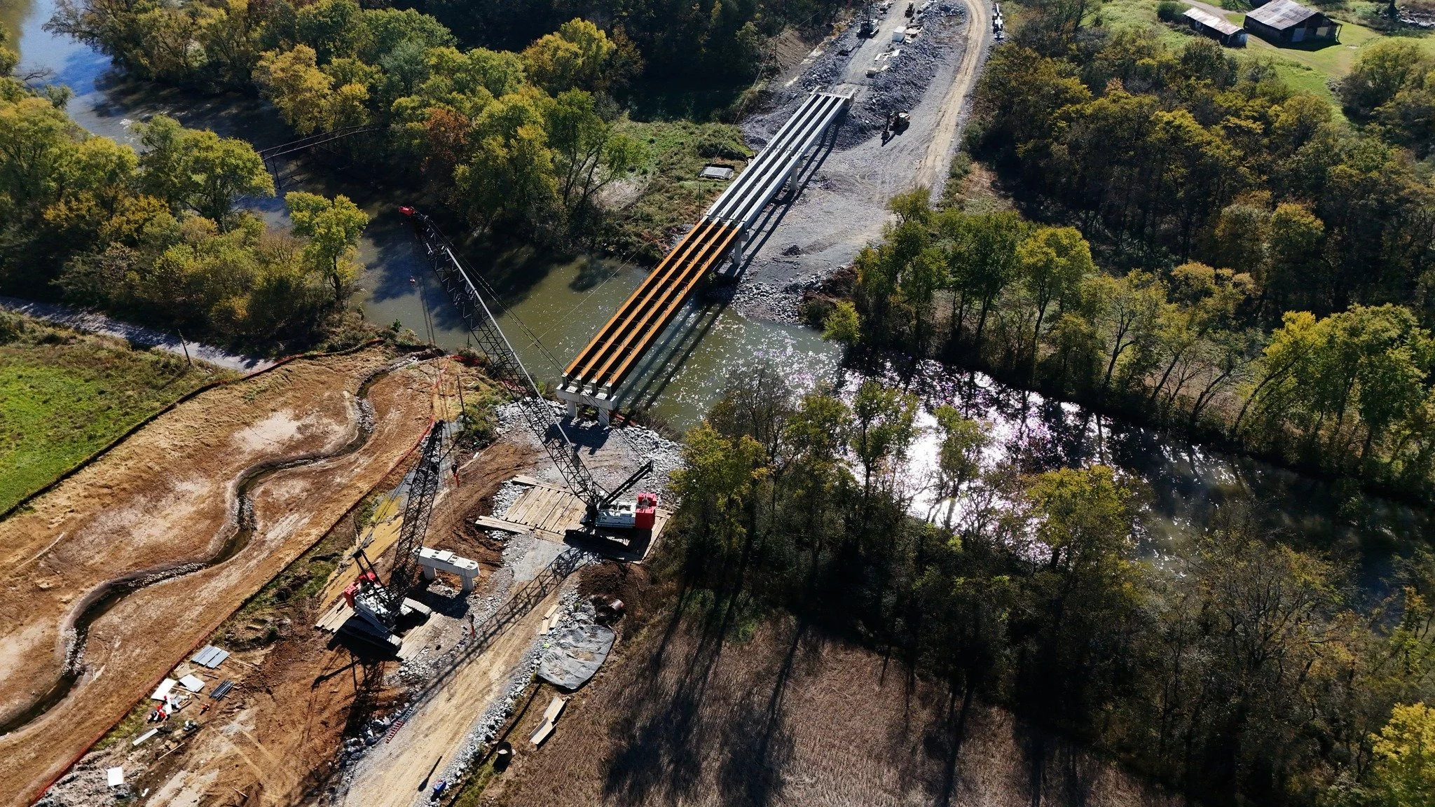 An aerial view of a bridge under construction over a river, surrounded by trees and construction equipment, with a dirt road leading to the site.