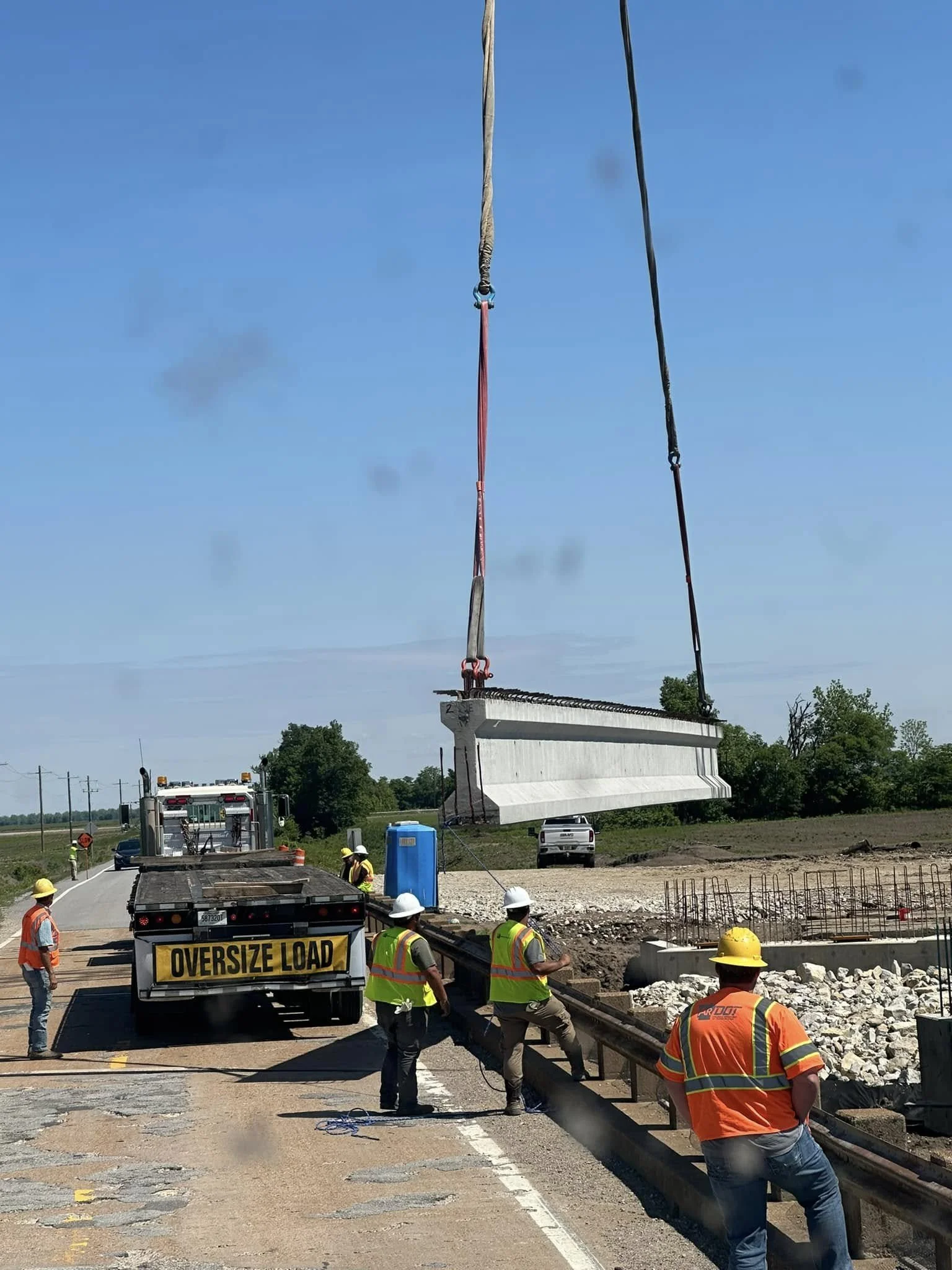 Construction workers on a road lifting a large concrete barrier using a crane with an oversize load truck, with a clear blue sky and trees in the background.