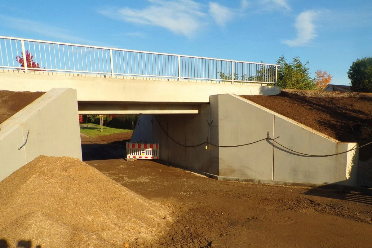 Underpass with concrete walls and metal railing above, dirt piles in the foreground, and trees with fall foliage in the background.