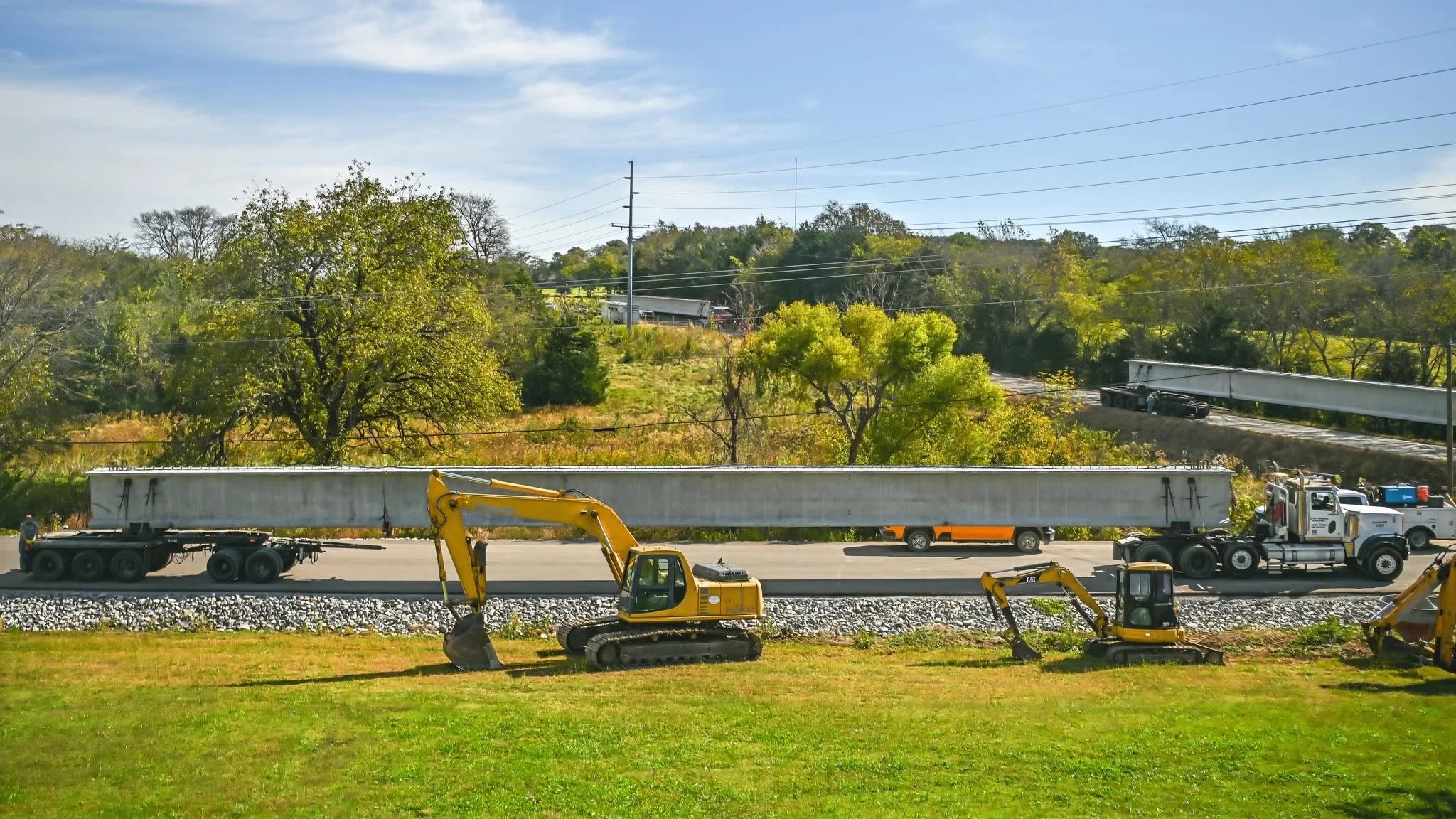 Construction site with excavators and trucks beside a railway track surrounded by greenery and trees under a cloudy sky.