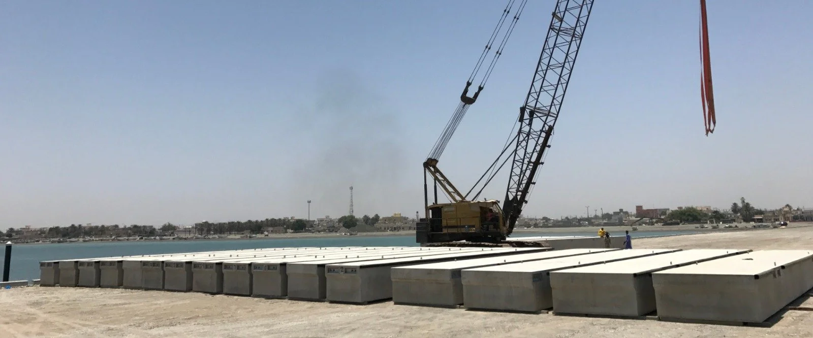 Construction site near a water body with a crane and large metal blocks being moved, and two workers in safety gear.