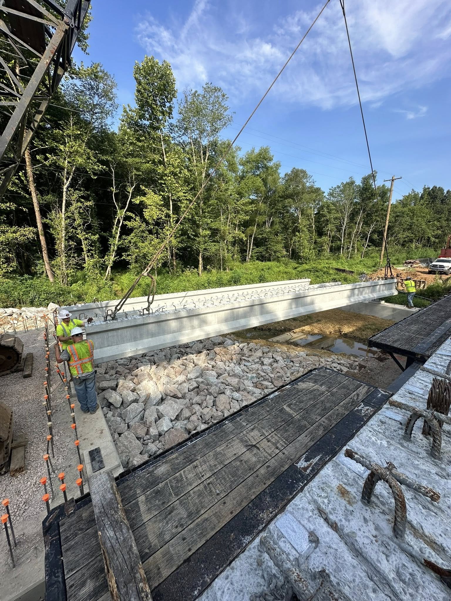 Construction workers installing a large steel beam at a bridge construction site, surrounded by rocks, wooden planks, and construction equipment, with a wooded area and blue sky in the background.