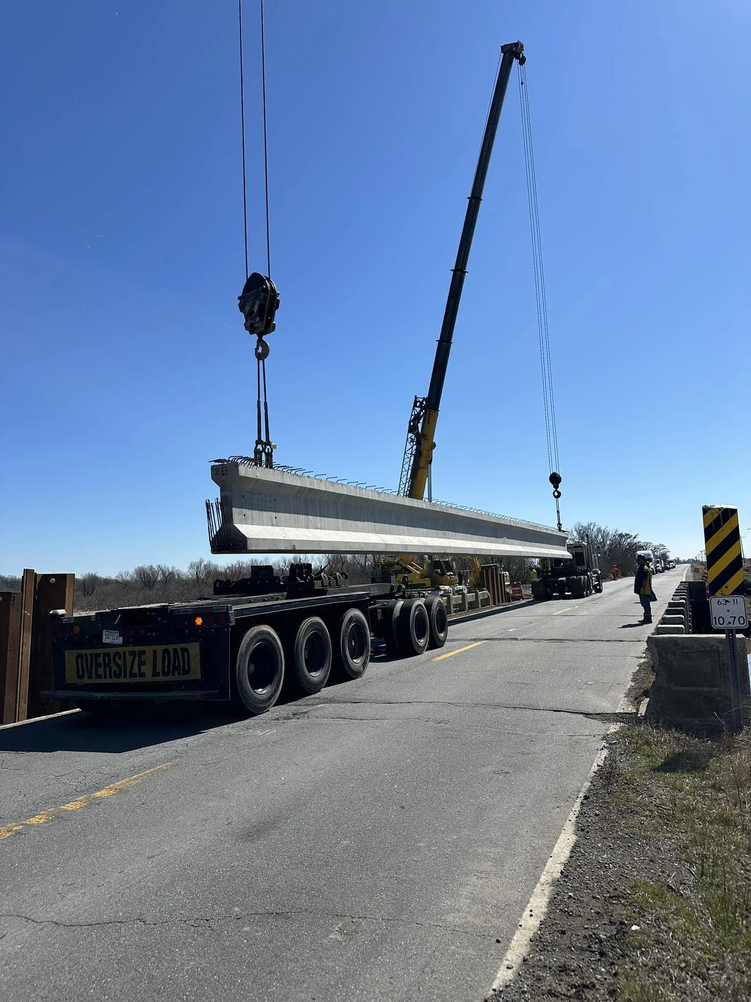 A large crane lifting a concrete bridge segment onto a road construction site, with workers overseeing the operation, and a sign indicating an oversized load.