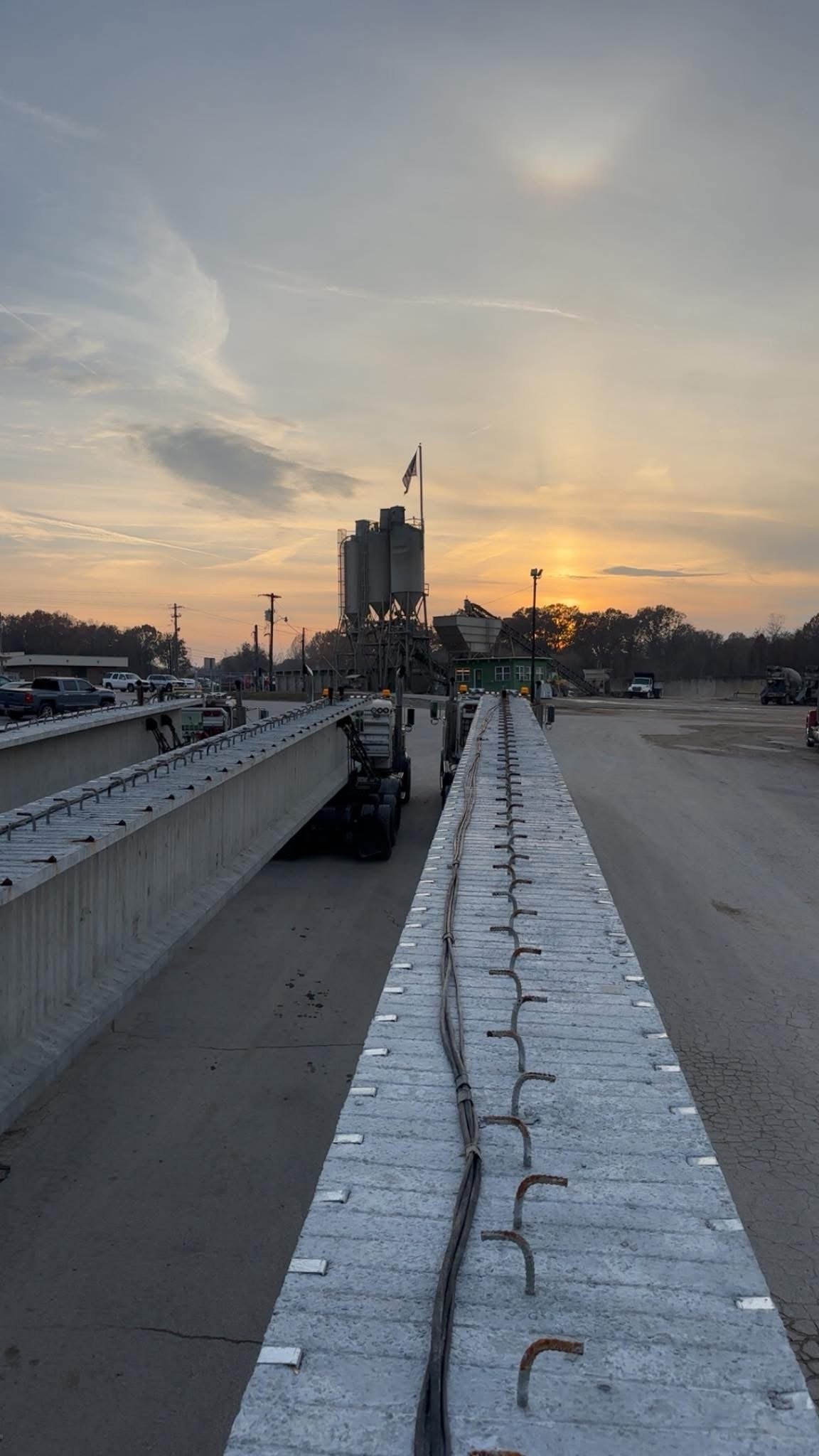 A construction site with concrete beams and steel rebar, equipment and vehicles parked around, sunset sky in the background.