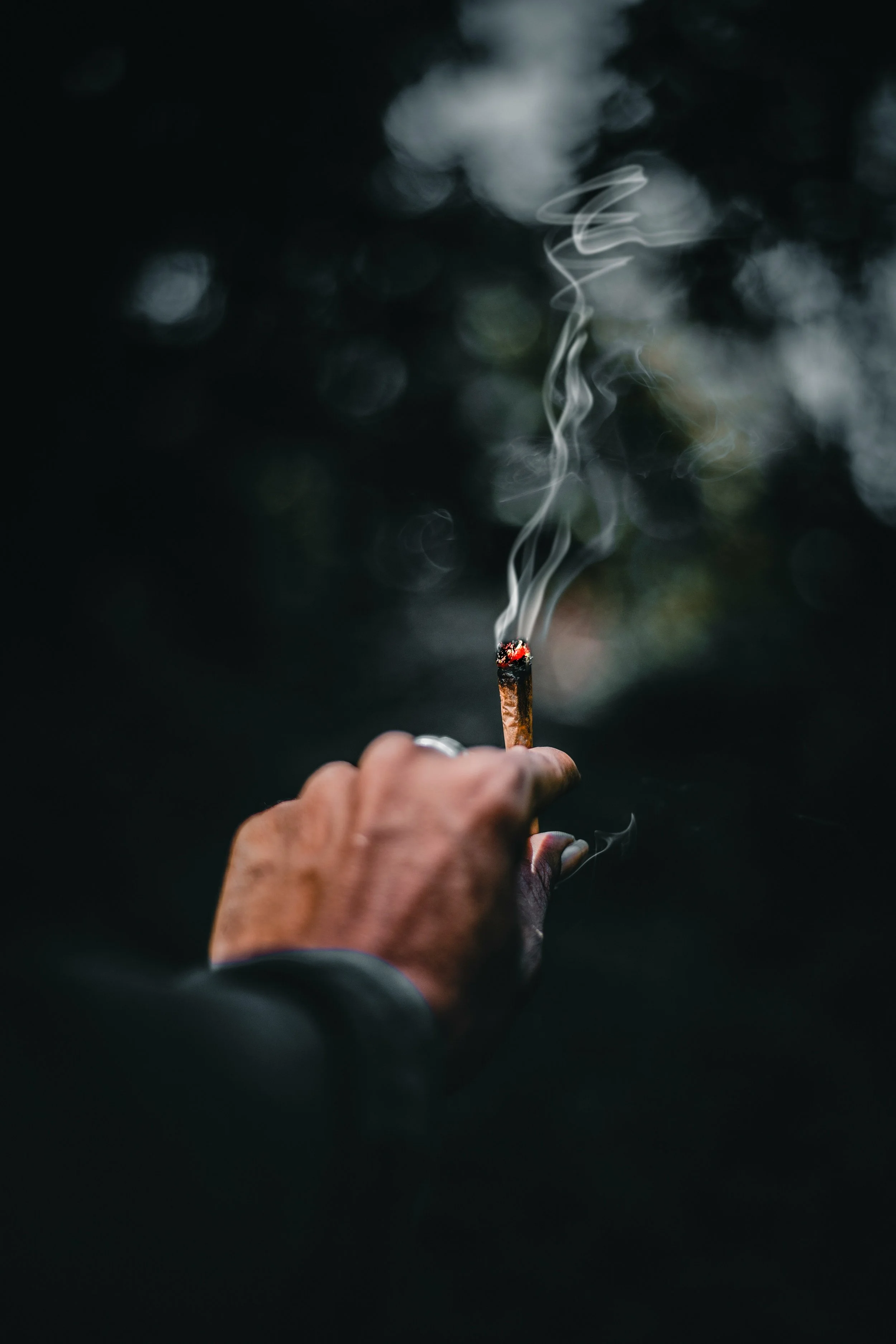 Close-up of a hand holding a cannabis joint, with smoke rising against a dark background.