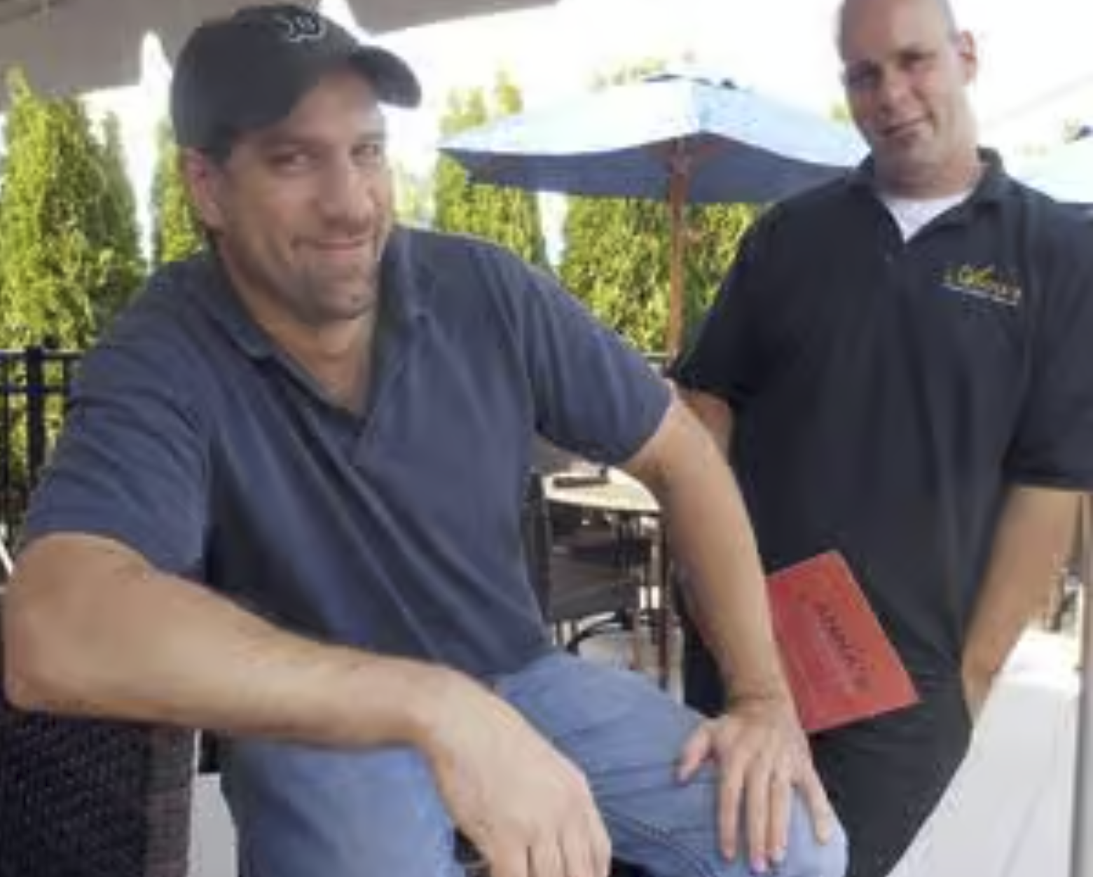 Two men are in an outdoor setting, one sitting and the other standing. The man sitting is wearing a dark baseball cap and a navy shirt, smiling, with his arm resting on his knee. The man standing is wearing a black polo shirt with a logo, has his right hand on the seated man's knee, and is holding a red menu or folder. There is a patio umbrella and greenery in the background.