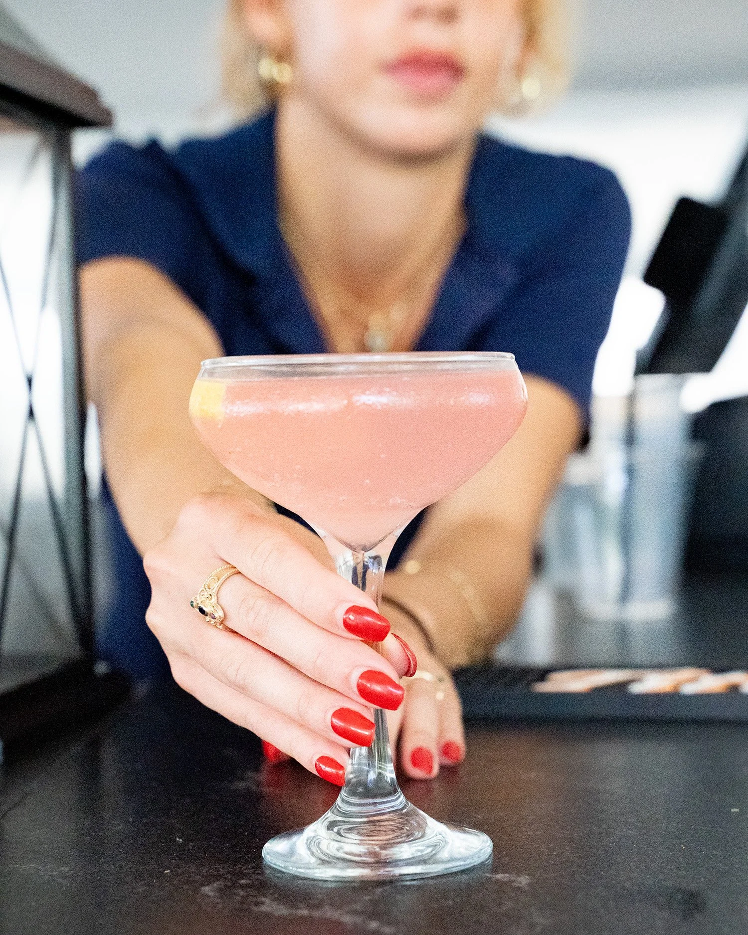 A woman with red nail polish and gold jewelry is holding a pink cocktail in a coupe glass, extending it toward the camera. She is wearing a navy blue top and is seated at a table in a setting that appears to be a bar or restaurant.