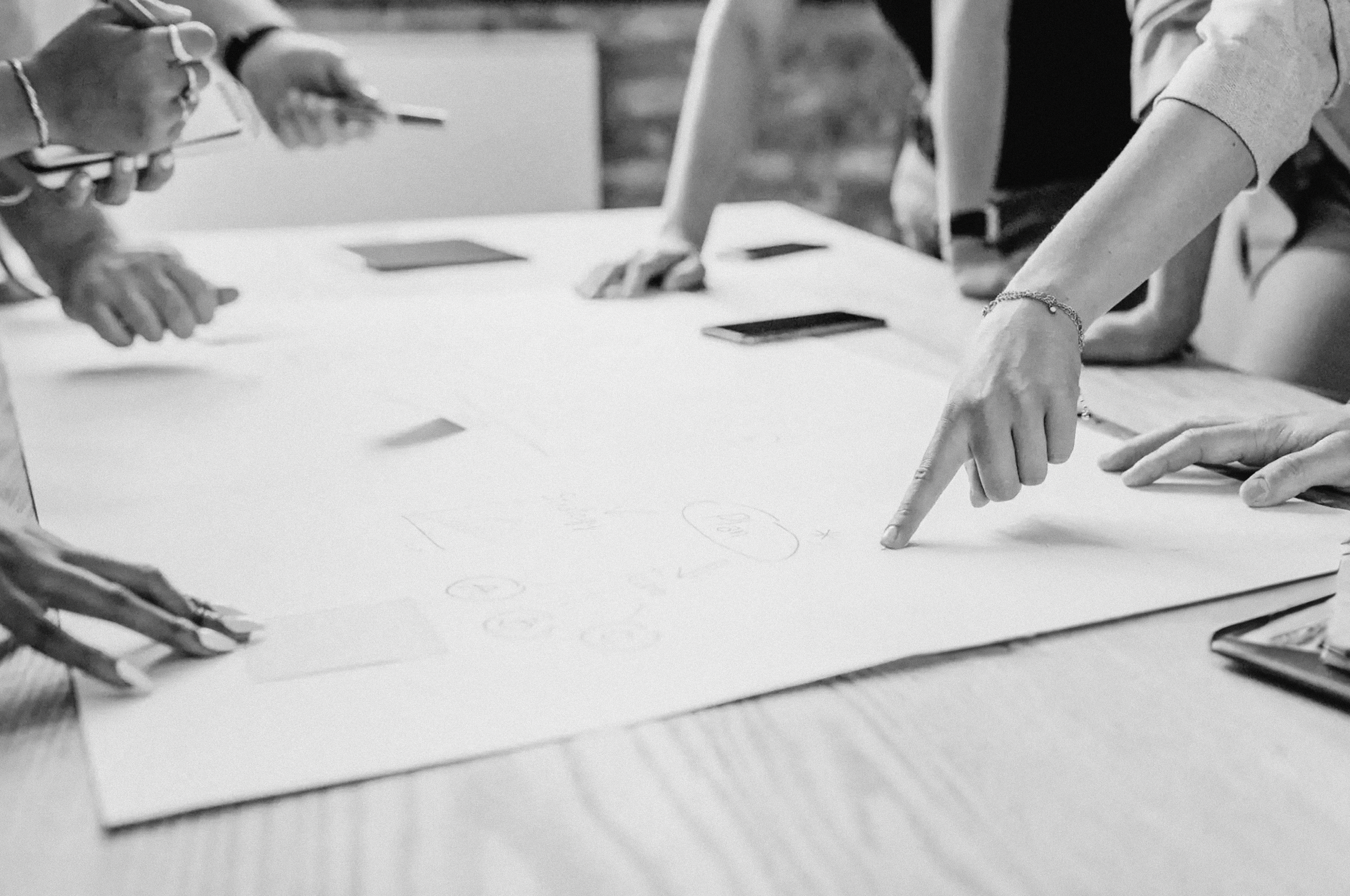 People gathered around a table, discussing and pointing at a large sheet of paper with sketches and notes, using smartphones.