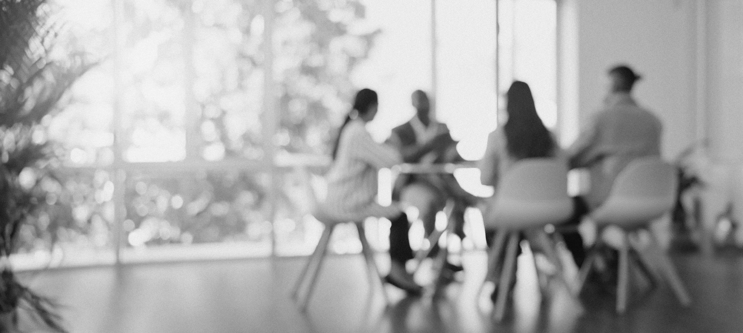 A blurred black-and-white photo of four people sitting around a table in a bright room with large windows, possibly engaged in a discussion or meeting.