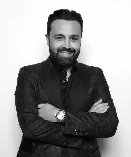 Black and white portrait of a man with a beard and mustache, wearing a dark blazer and shirt, smiling with arms crossed, against a plain background.