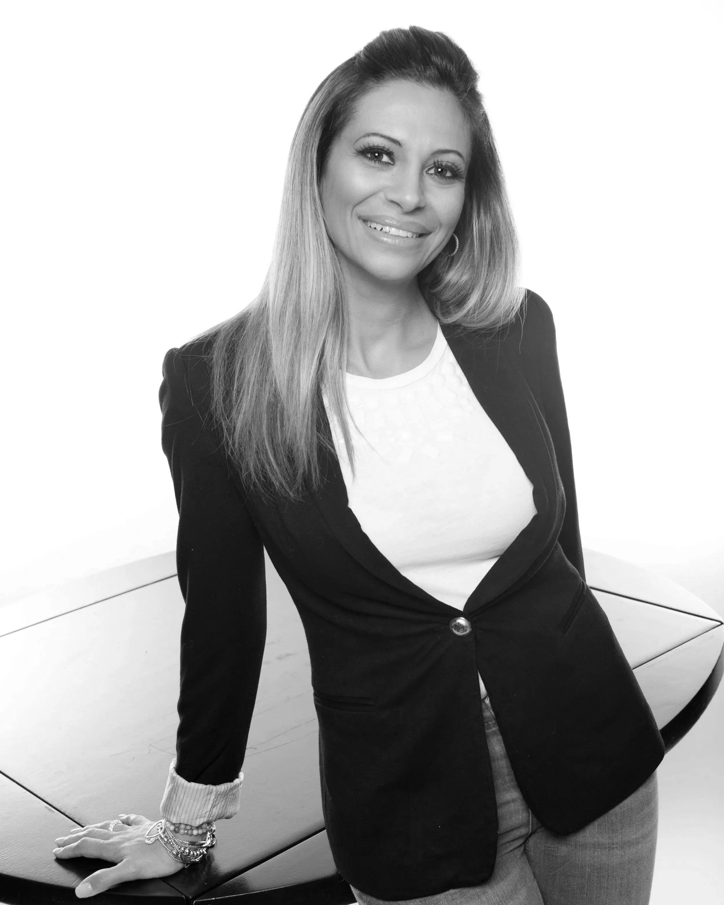 A woman with shoulder-length hair smiling, wearing a blazer over a white top, posing with her hand on a table.