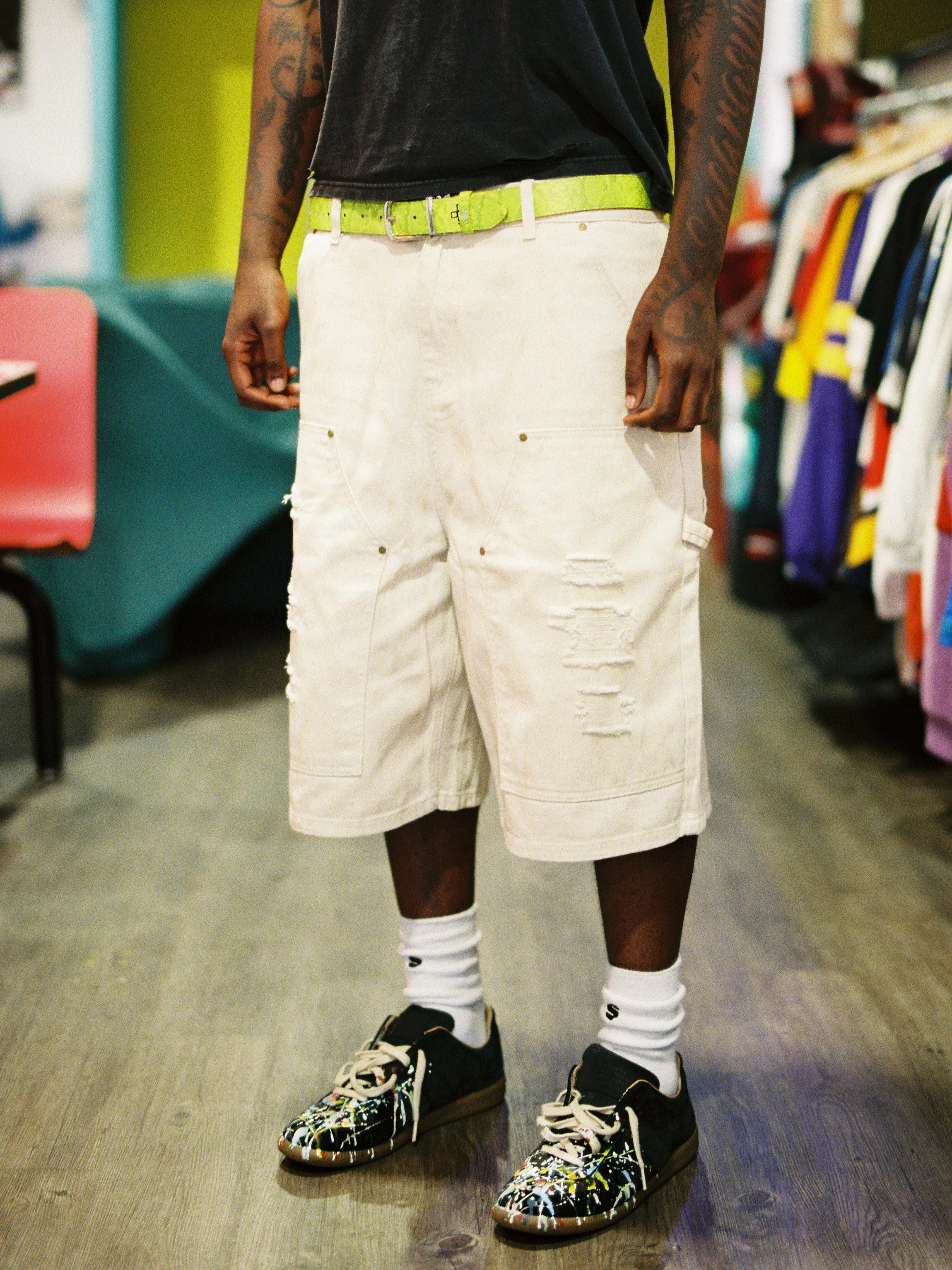 A person wearing black sneakers with a colorful splattered paint design, white crew socks, white distressed shorts, a bright yellow belt, and a black t-shirt, standing on a wooden floor in a retail store with clothing racks in the background.