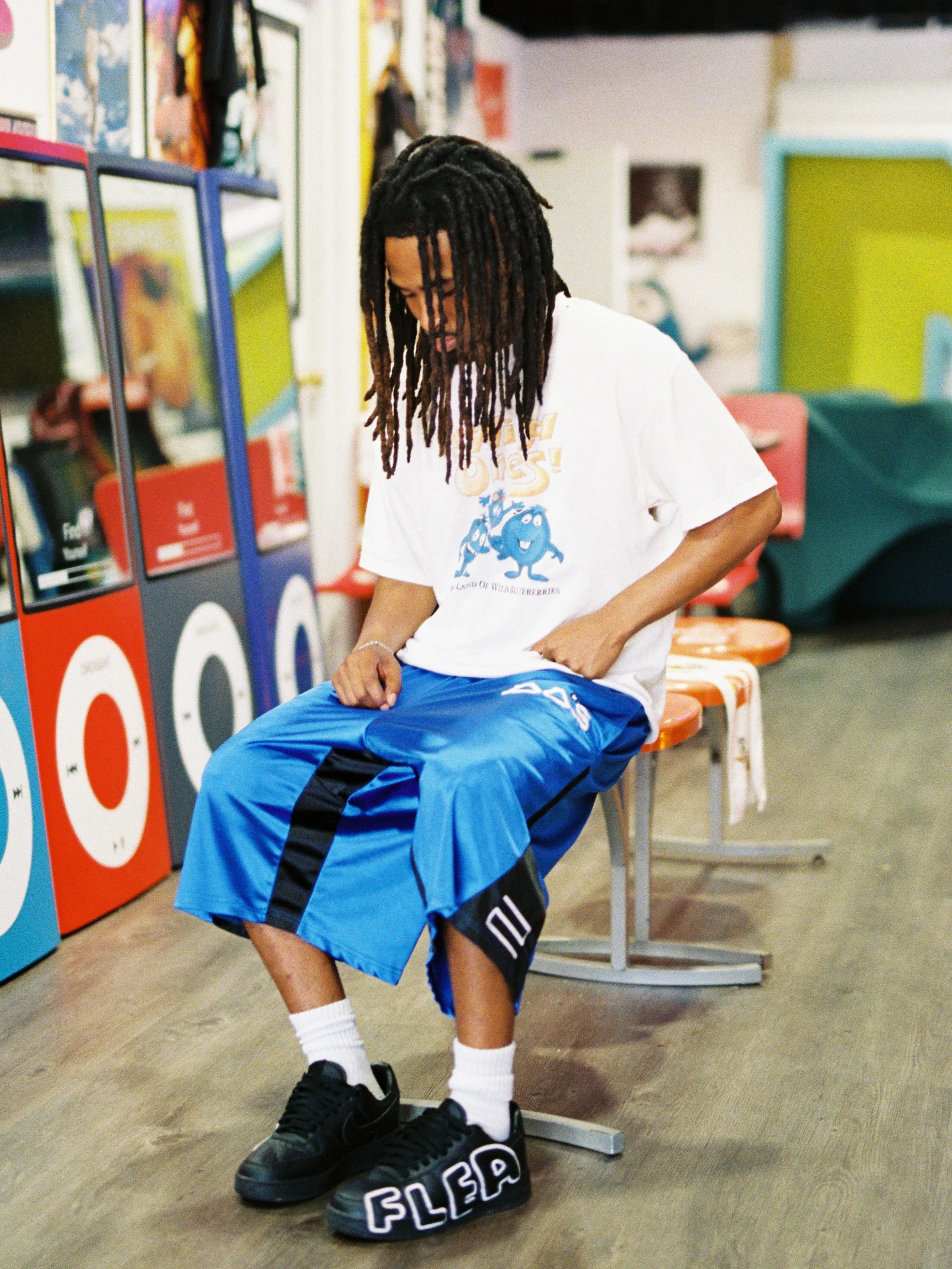 A young man with dreadlocks sits on a stool at an indoor space that appears to be a store or arcade. He wears a white T-shirt with a graphic, blue baggy shorts, white athletic socks, and black sneakers. His eyes are closed or looking down, and his ri