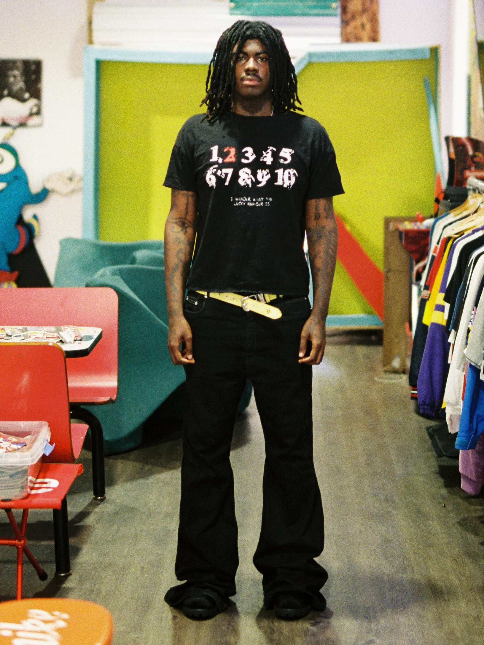A young man with dreadlocks wearing a black t-shirt with numbers and text, black pants, and large black shoes standing in a colorful indoor space with clothing racks and chairs around him.