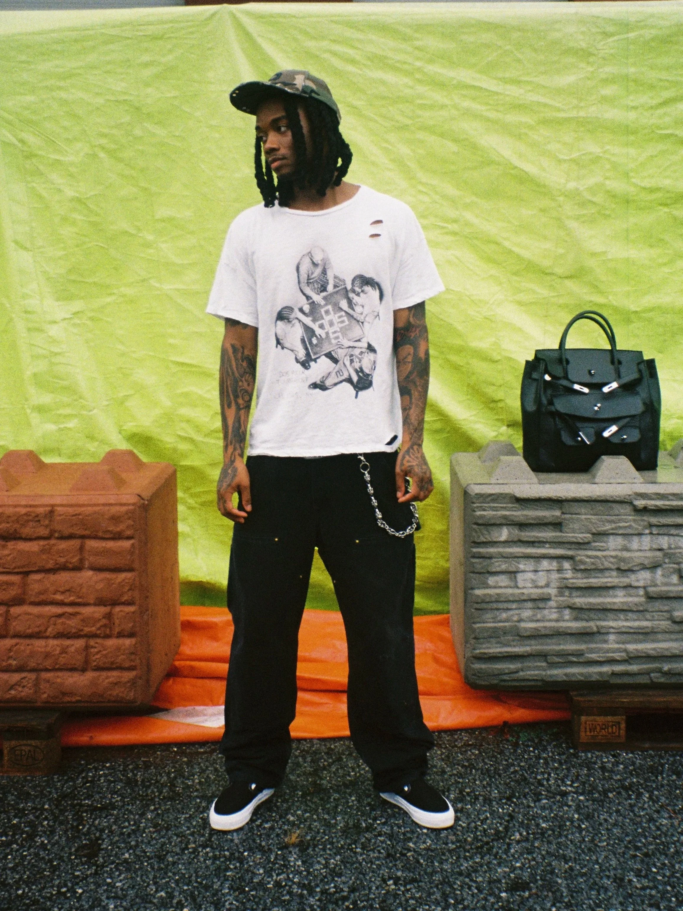 A young man with dreadlocks, wearing a black camouflage cap, a white distressed T-shirt featuring graffiti-style artwork, black pants with chain accessories, and black and white sneakers, standing in front of a green backdrop with brick-like structur