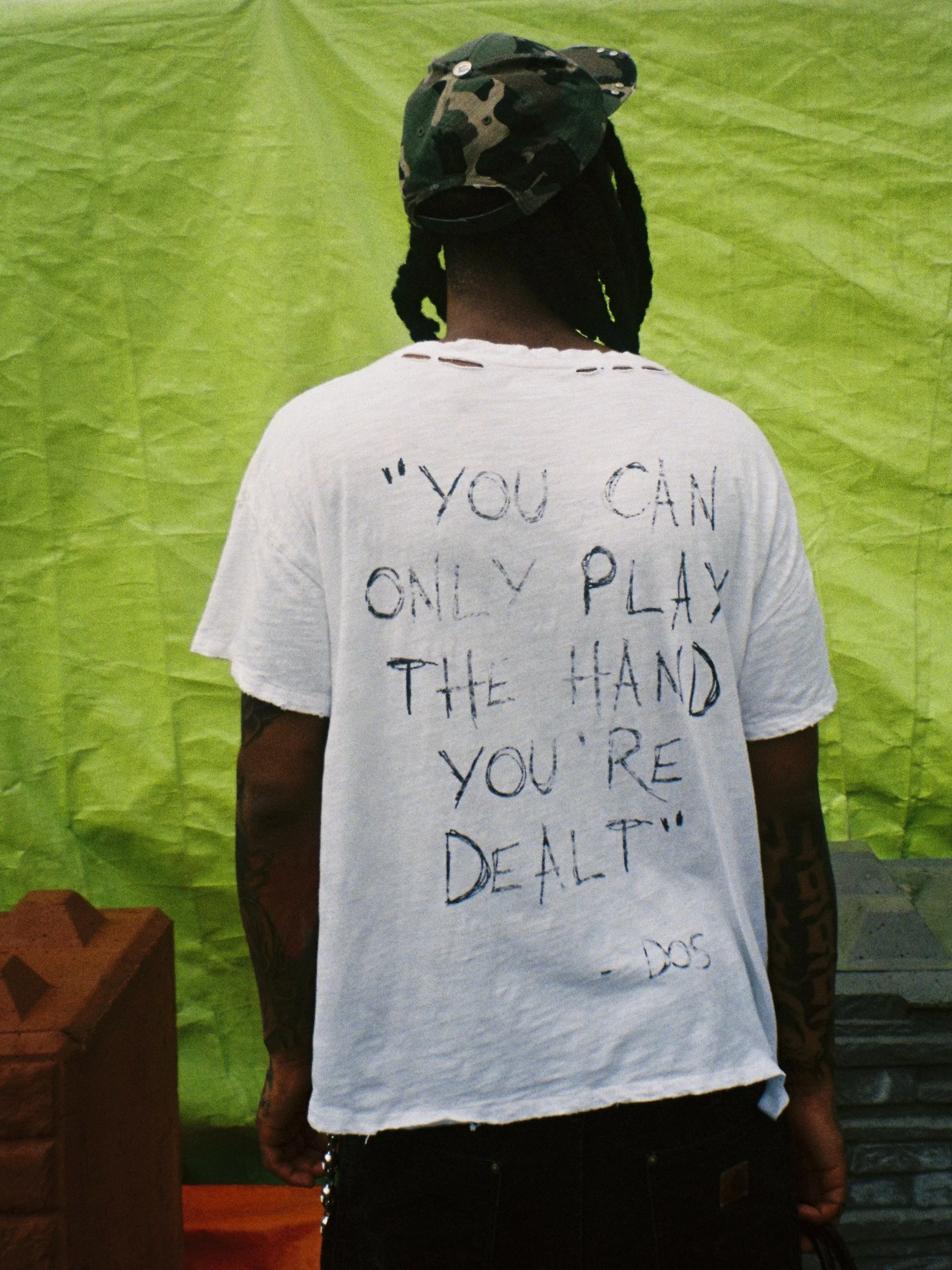 Back of a person wearing a white T-shirt with handwritten text, a camouflage cap, and tattoos on their arm, standing in front of a green background.
