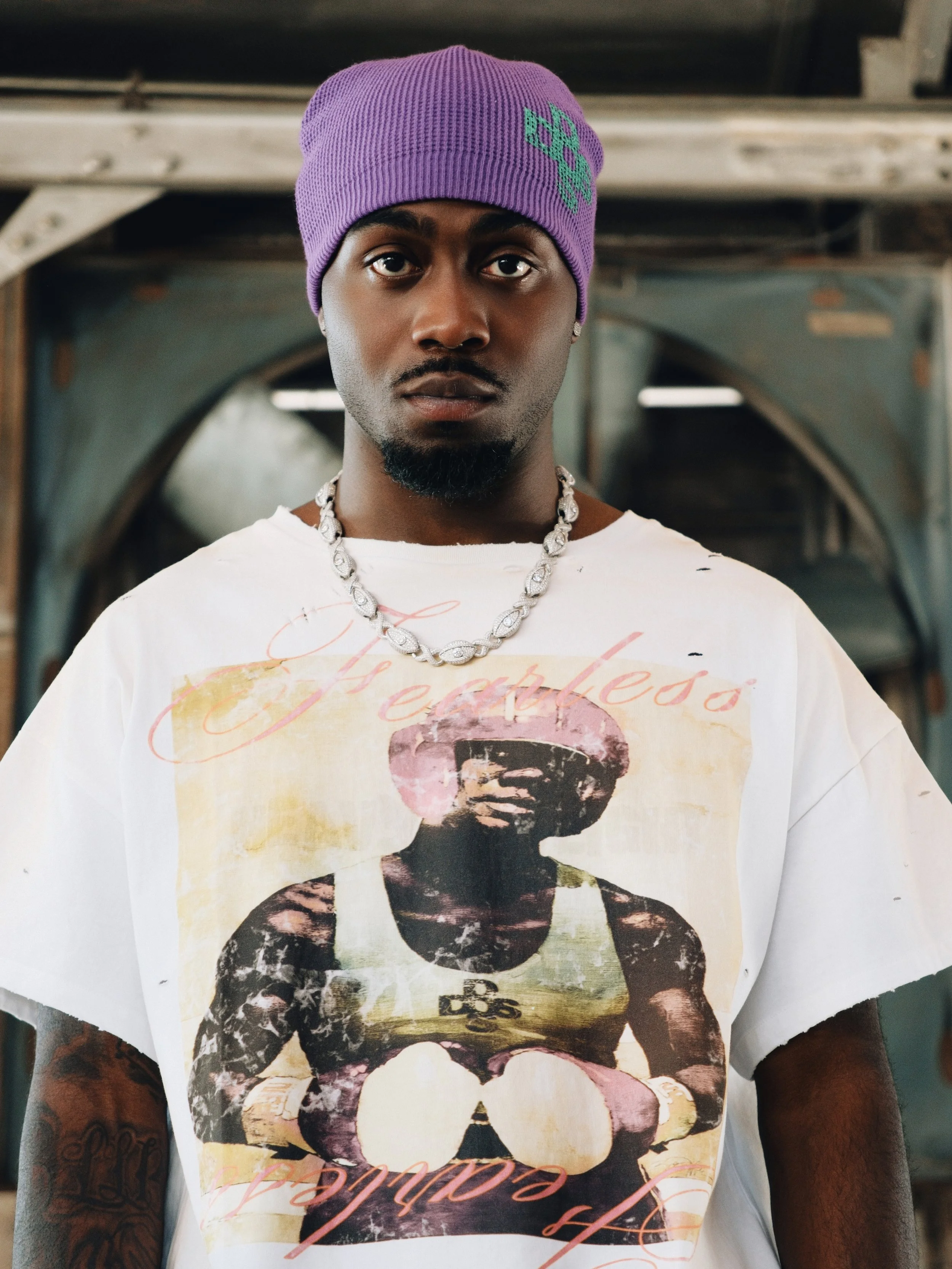A young Black man wearing a purple beanie, white graphic T-shirt, and a silver chain necklace, standing in an industrial or warehouse setting with metal structures in the background.