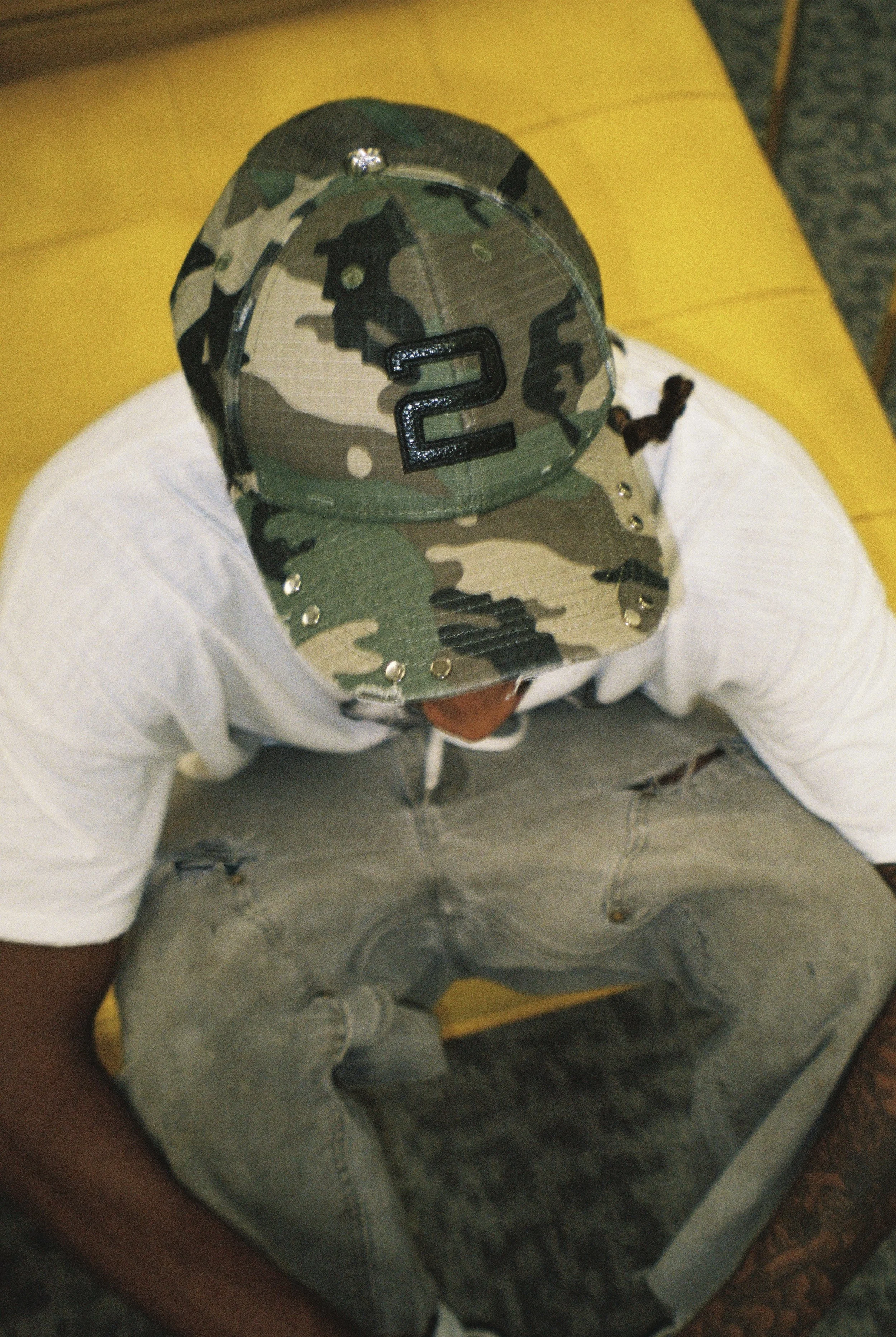 A person wearing a camouflage baseball cap with a large black number 2 on the front, white shirt, and light gray shorts, sitting on a yellow chair.