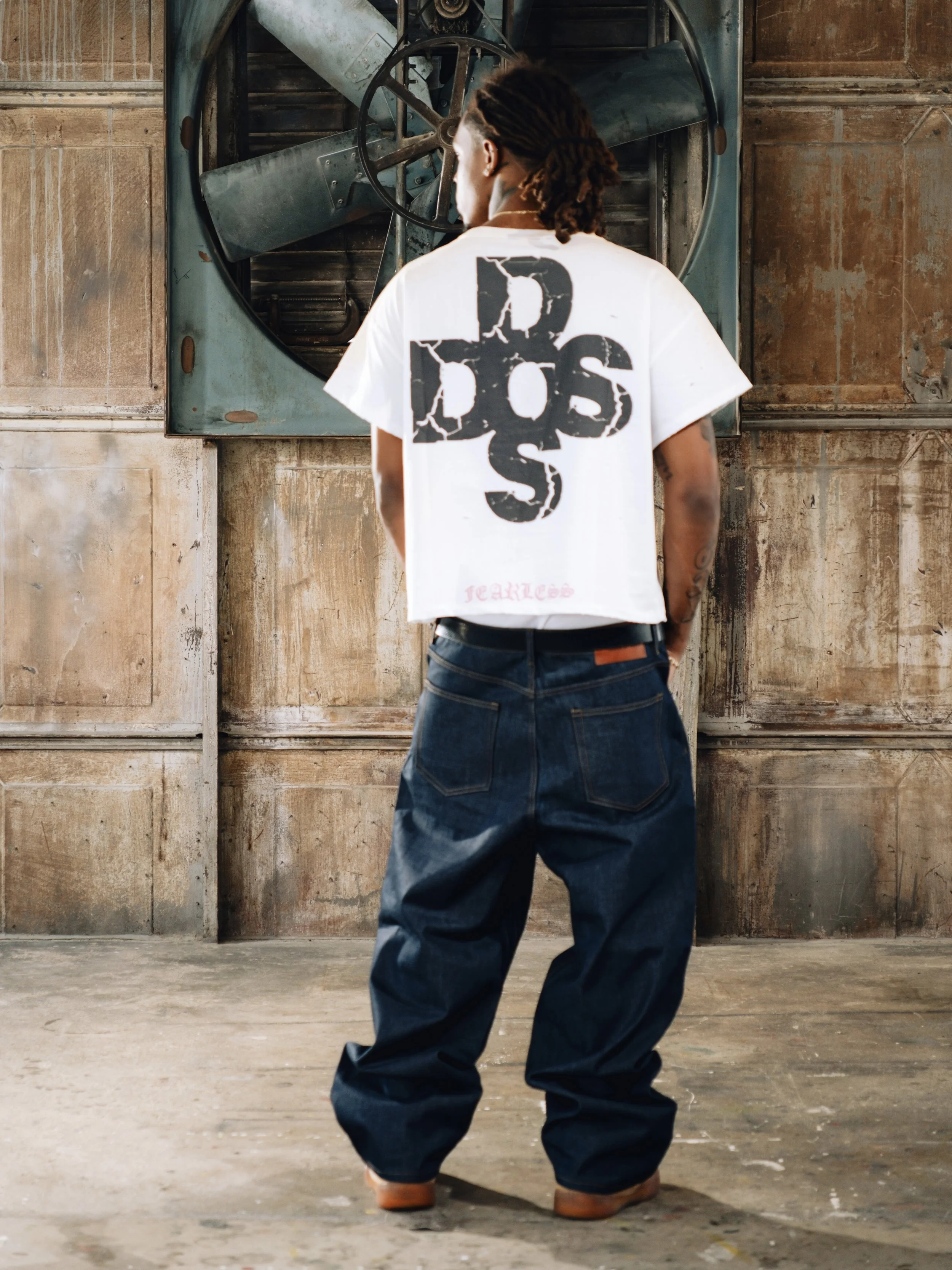 Person with dreadlocks wearing a white T-shirt and baggy jeans, standing in an industrial space with a large metal fan on the wall.