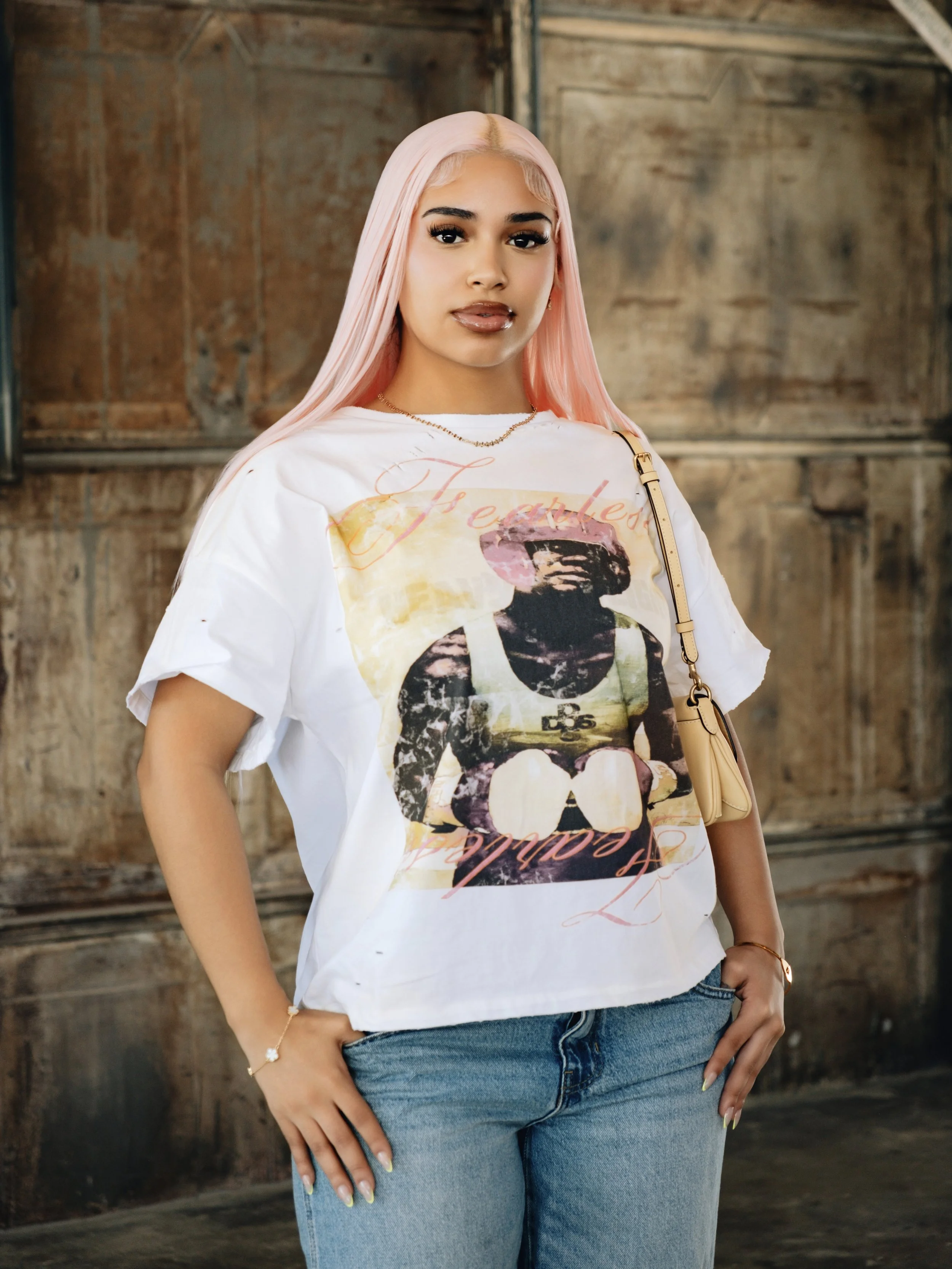 A woman with long pink hair wearing a white graphic T-shirt and blue jeans, standing in front of a rustic wooden wall.