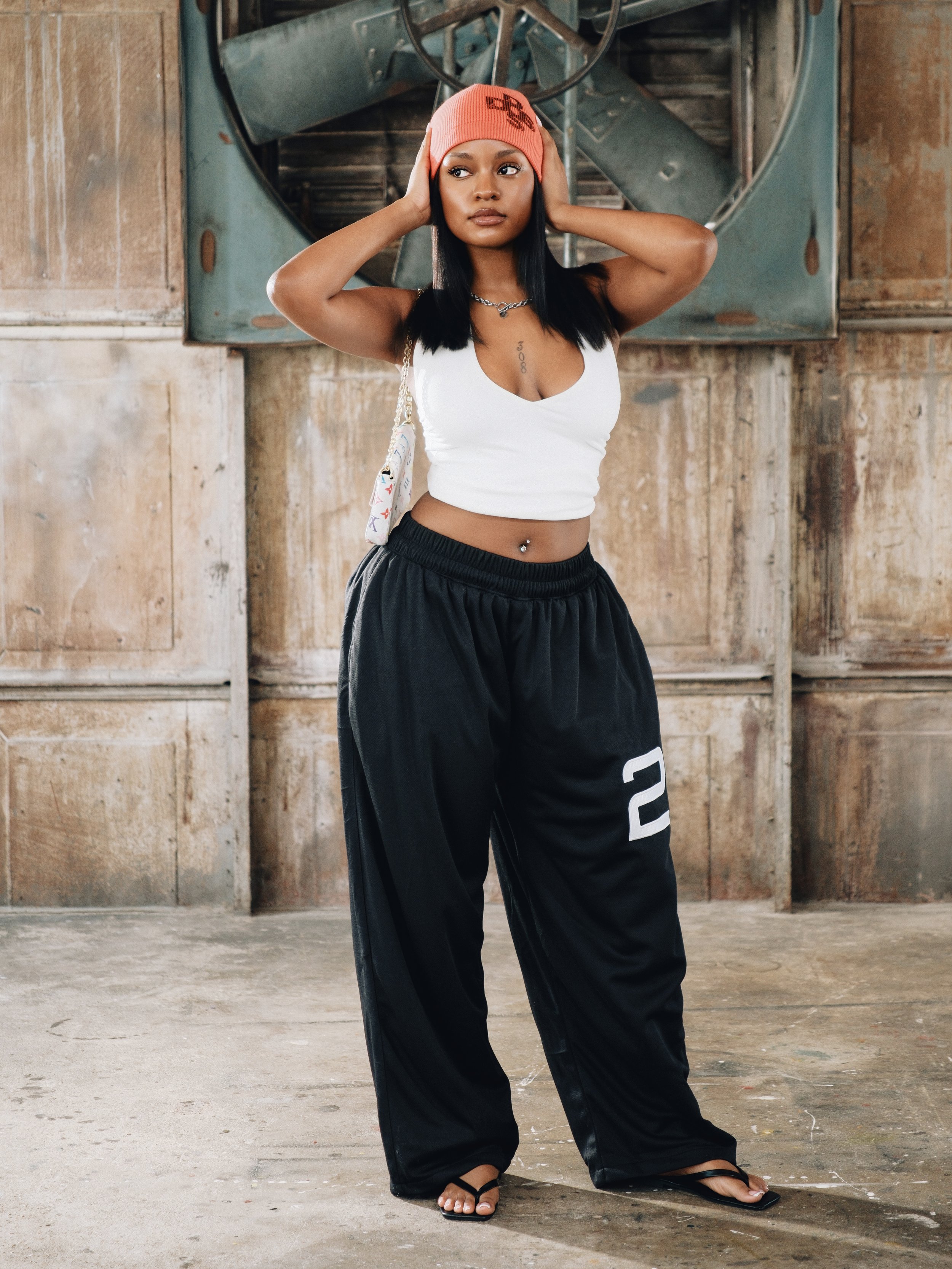 A woman with medium skin tone wearing a red beanie, white crop top, black baggy pants with a number 2 on the leg, black sandals, and a silver chain necklace, standing in an industrial setting with a large metal fan and concrete walls.