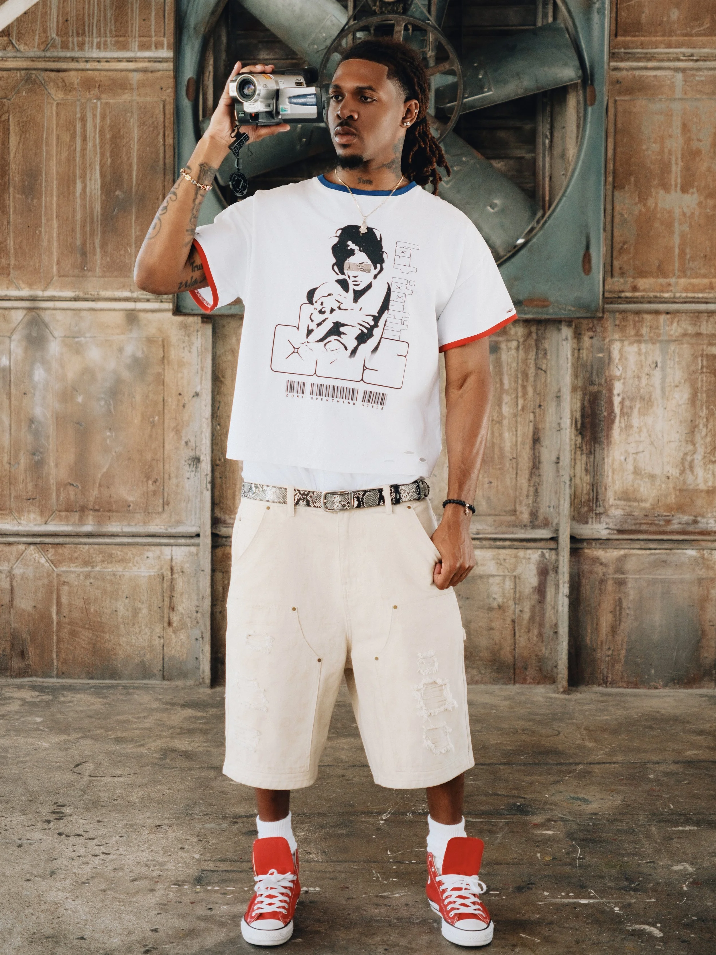 A man with dreadlocks and tattoos wearing a graphic t-shirt, beige ripped shorts, red sneakers, and white socks standing in an industrial setting, holding a video camera to his head.
