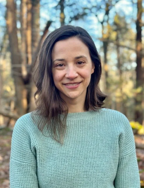A woman with shoulder-length brown hair smiling outdoors in a wooded area during daytime.