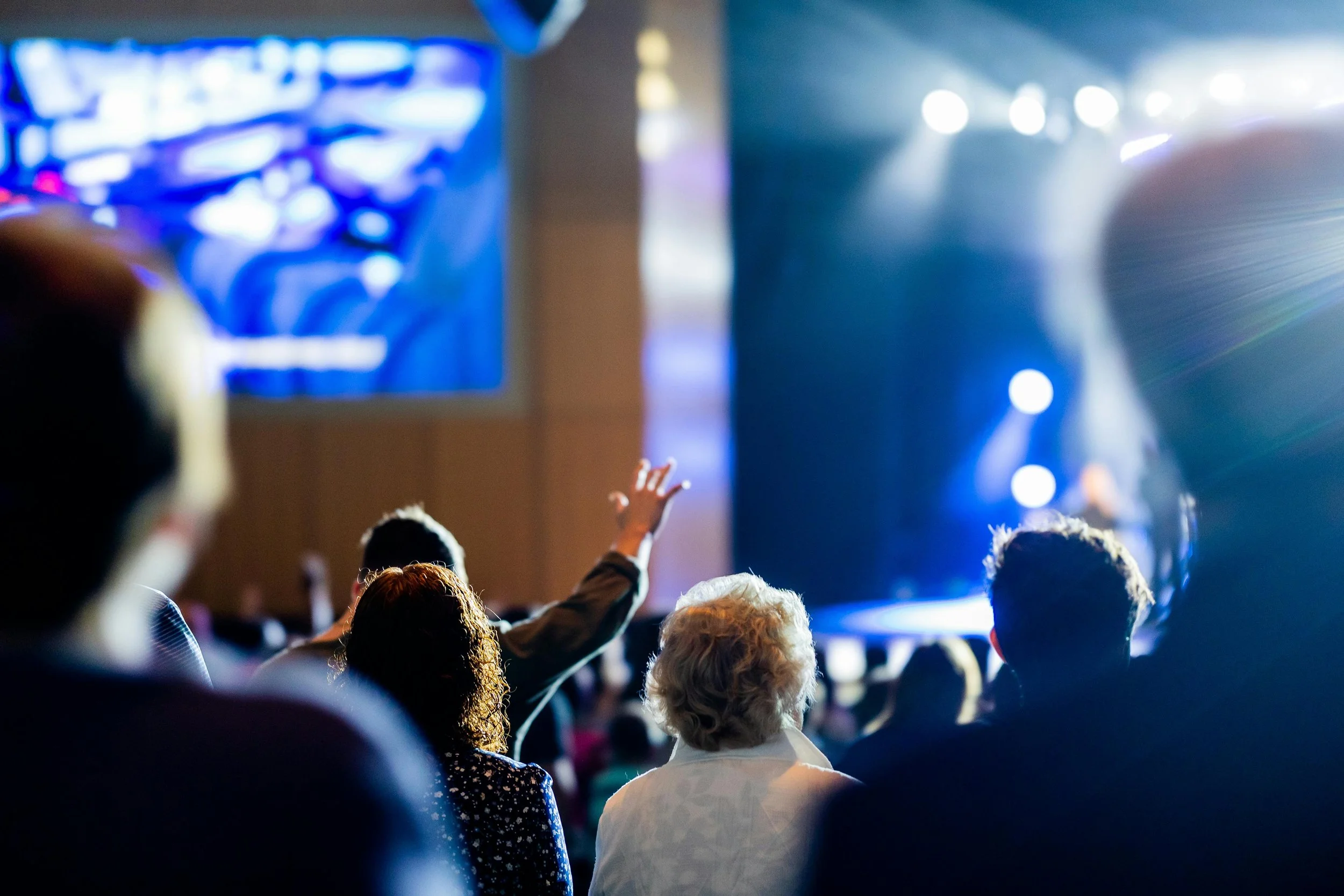 Audience members attending a concert or event, some raising their hands, seated in front of a brightly lit stage with colorful lights.