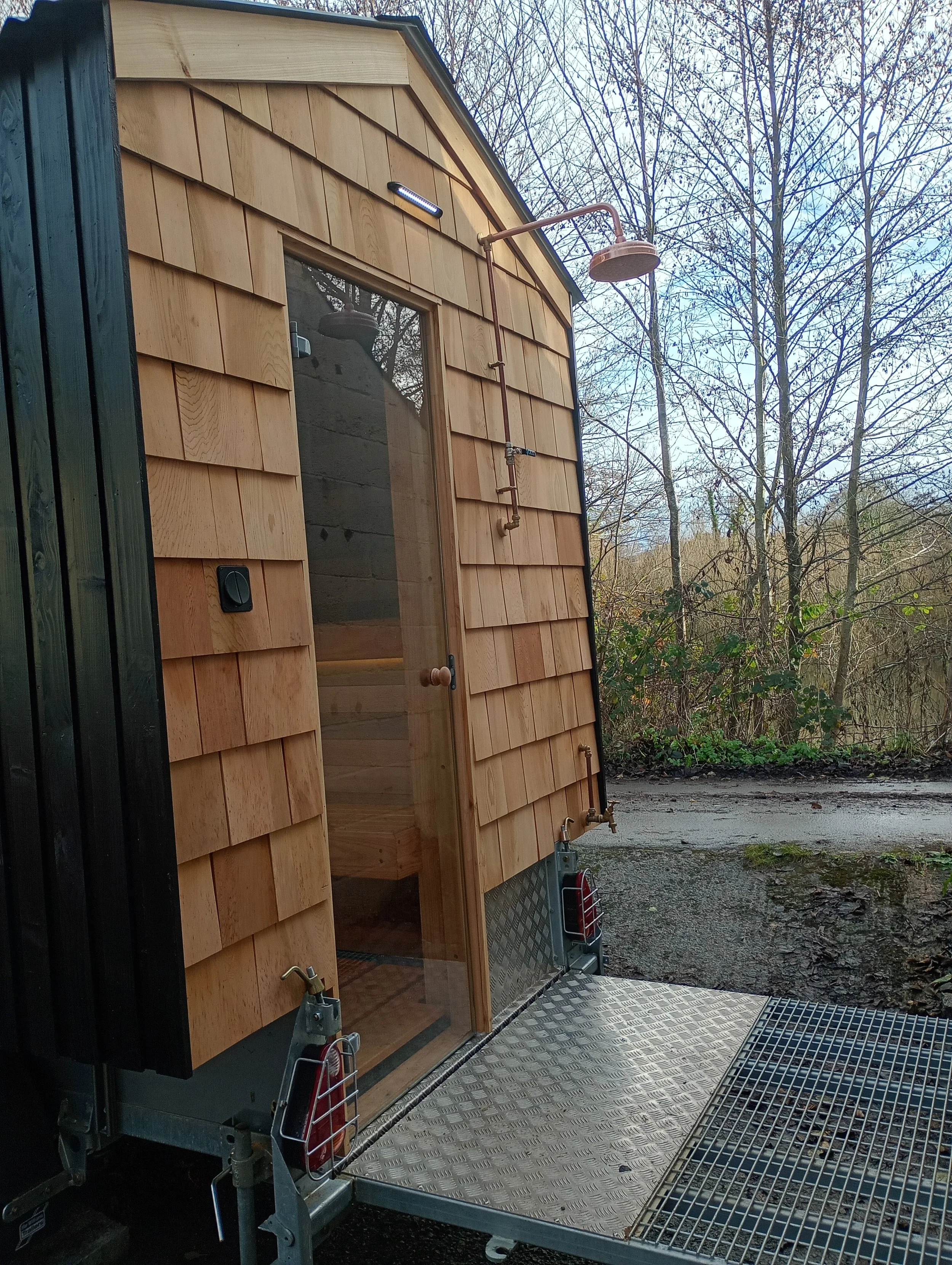 Wooden tiny house with outdoor shower, glass door, and metal platform, set against trees and sky.